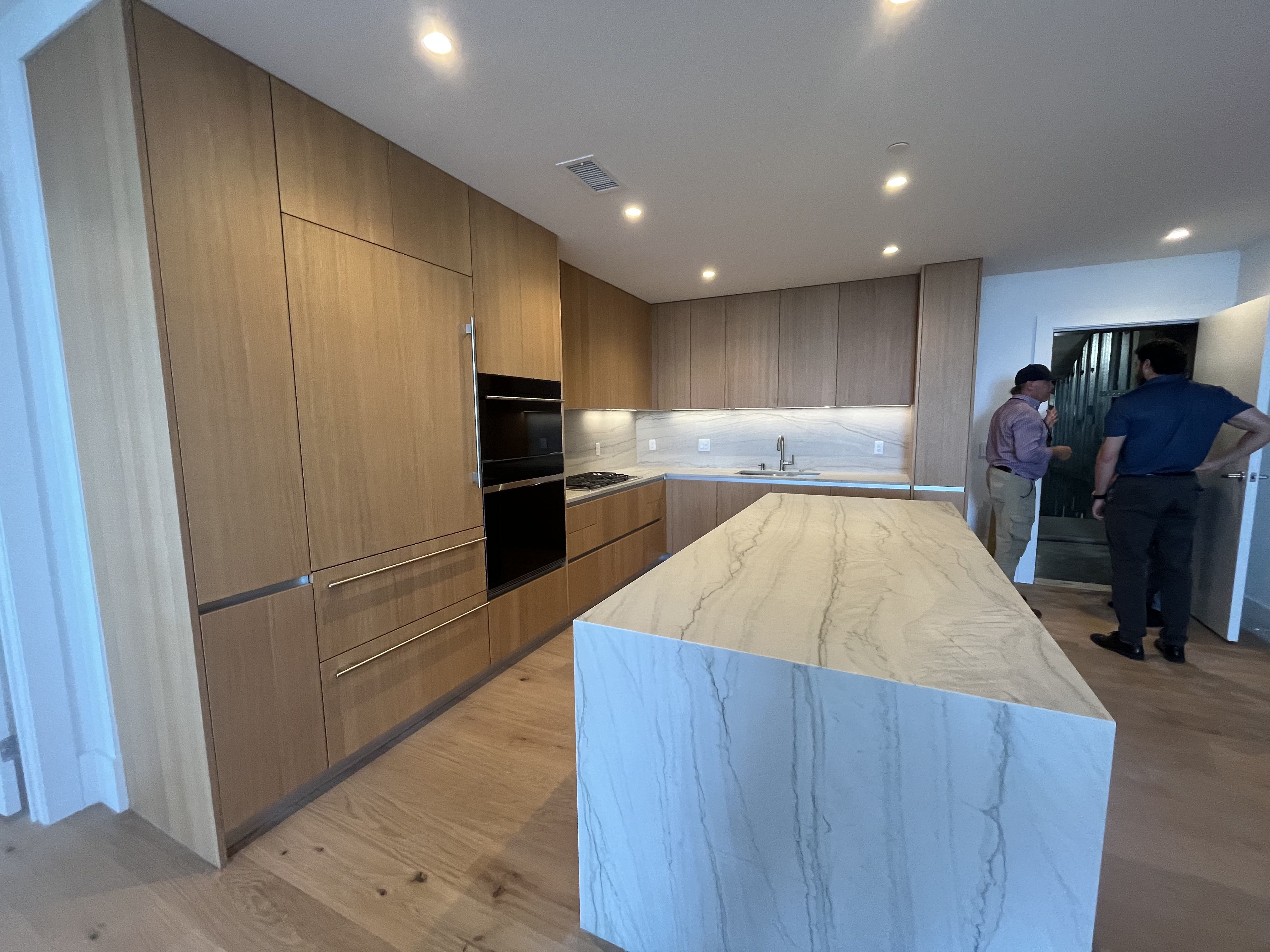 Modern kitchen with tall light brown wood cabinets along two walls, a marble island and white-veined countertops, built-in oven, and recessed ceiling lights. Two people stand near a doorway in the background.