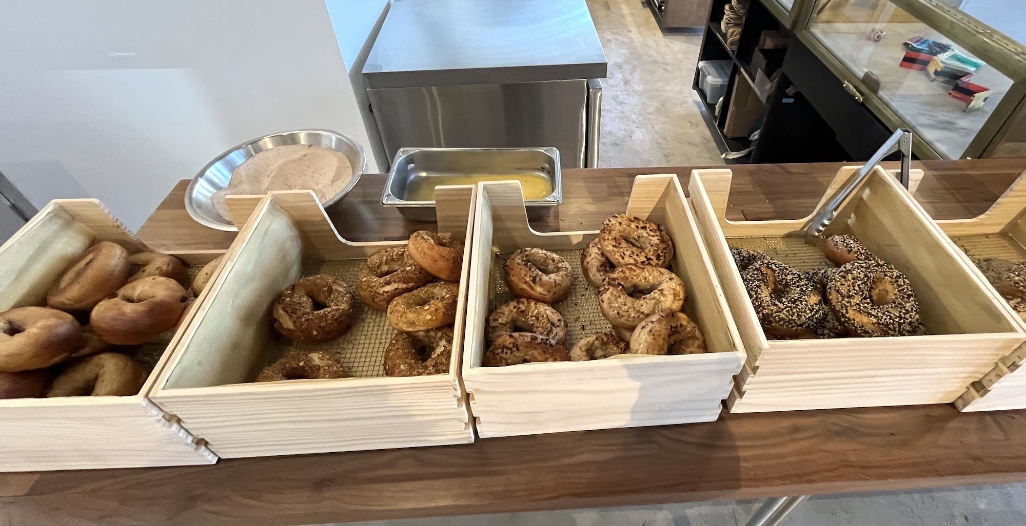 Wooden trays holding various bagels, some plain and others topped with seeds, arranged on a wooden counter with plates and utensils nearby in a bakery setting.