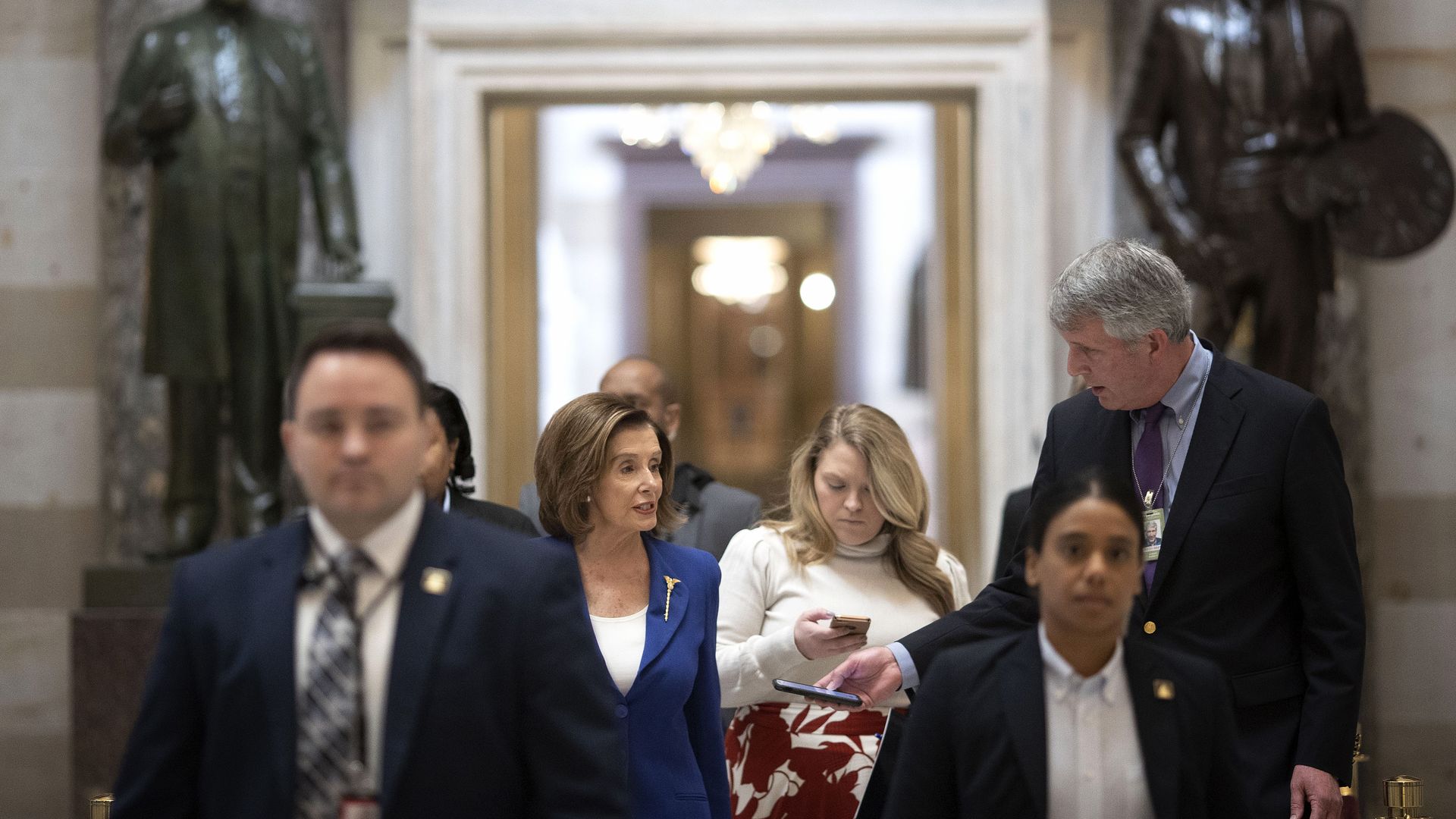 Photo of Nancy Pelosi and security guards/reporters walking through the halls of the U.S. Capitol