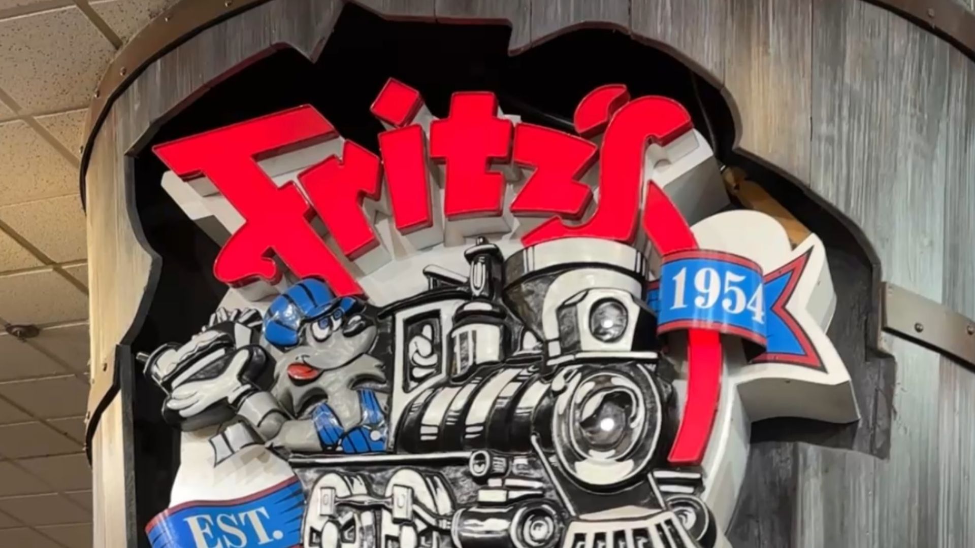 Close-up of a circular mural featuring bold red 3D lettering at the top, a black-and-white steam locomotive below, and a blue ribbon marked 1954 on the right.