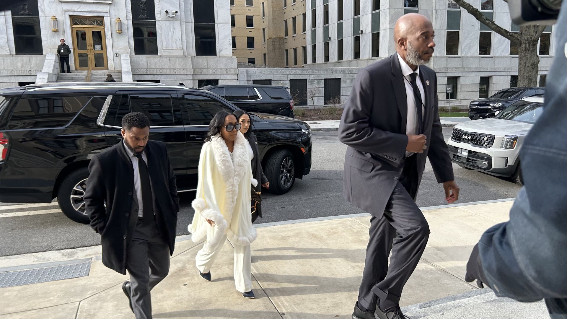 Group of four adults, three men and one woman in a white fur-trimmed coat and sunglasses, walking on a sidewalk near black vehicles and a building with marble facade.