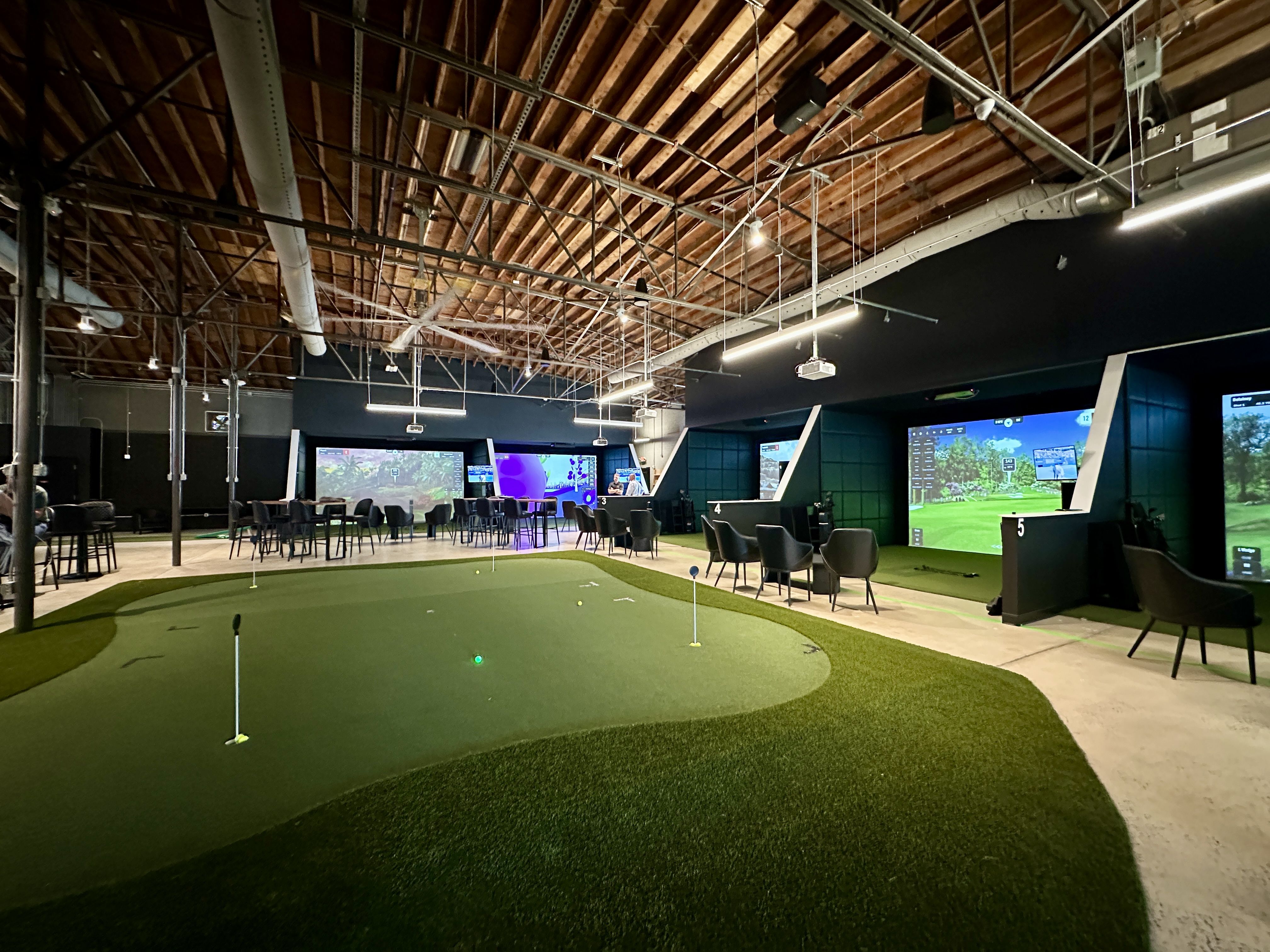 Indoor golf simulation center with artificial putting green in foreground, multiple golf simulator bays with large screens and seating, exposed wooden ceiling, and modern lighting.