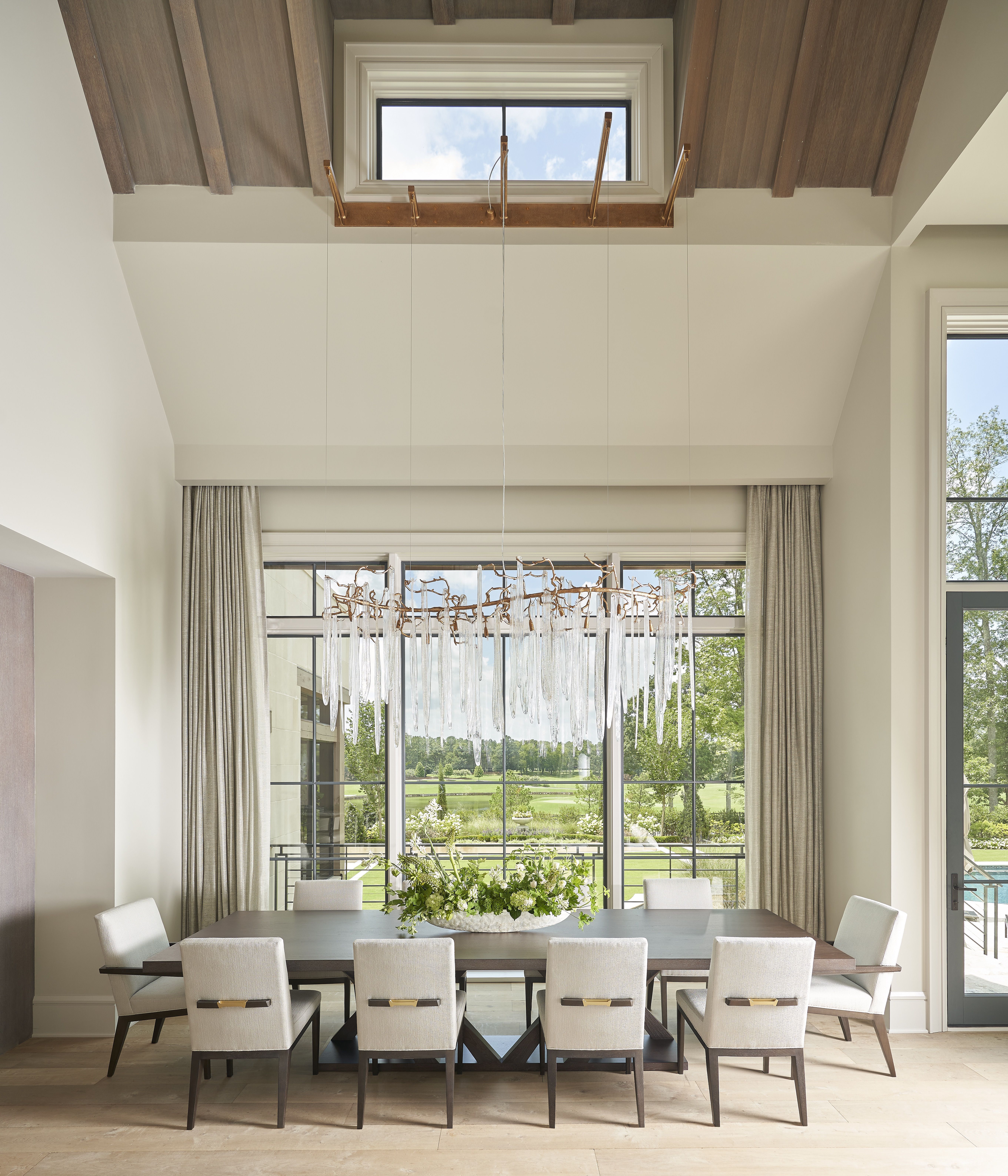Bright dining room with a long dark wood table, white upholstered chairs, and a crystal chandelier with dangling prisms. Large windows reveal a green lawn; beige curtains frame the scene.