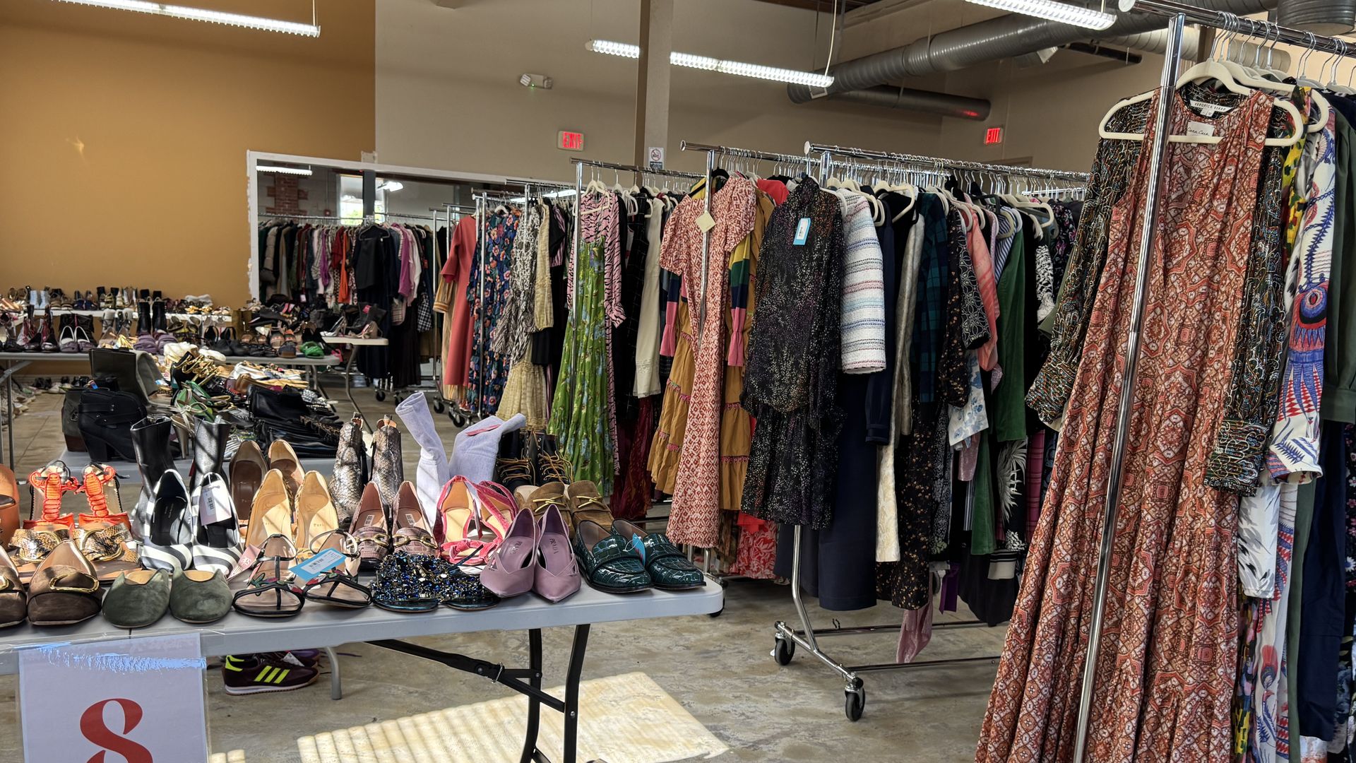 Indoor thrift or consignment store with racks of colorful dresses and tables filled with various shoes like heels, sandals, and boots under fluorescent lights.