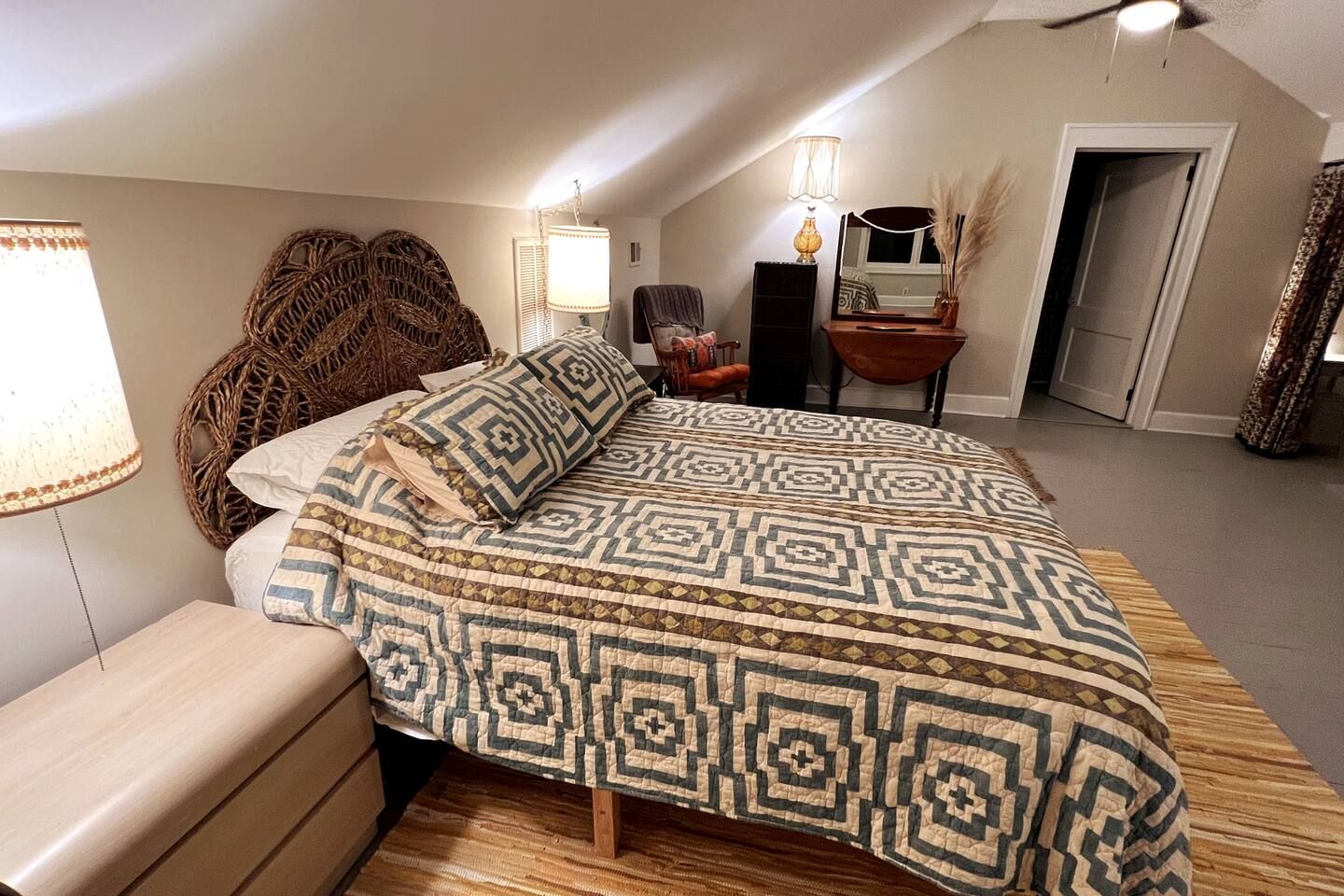 Cozy bedroom with a large bed featuring a geometric patterned quilt in blue, beige, and brown, wicker headboard, wooden nightstand, two lamps, a chair, dresser, and a vanity with pampas grass.