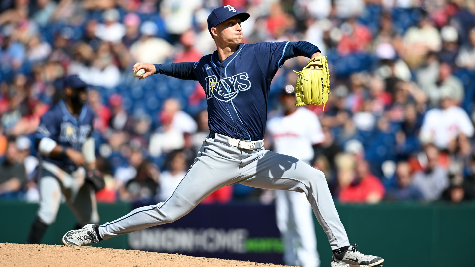 A baseball pitcher wearing a dark blue jersey and hat, light gray pants and a yellow glove on his left hand winds up to throw a pitch, with fans seated in the background. 
