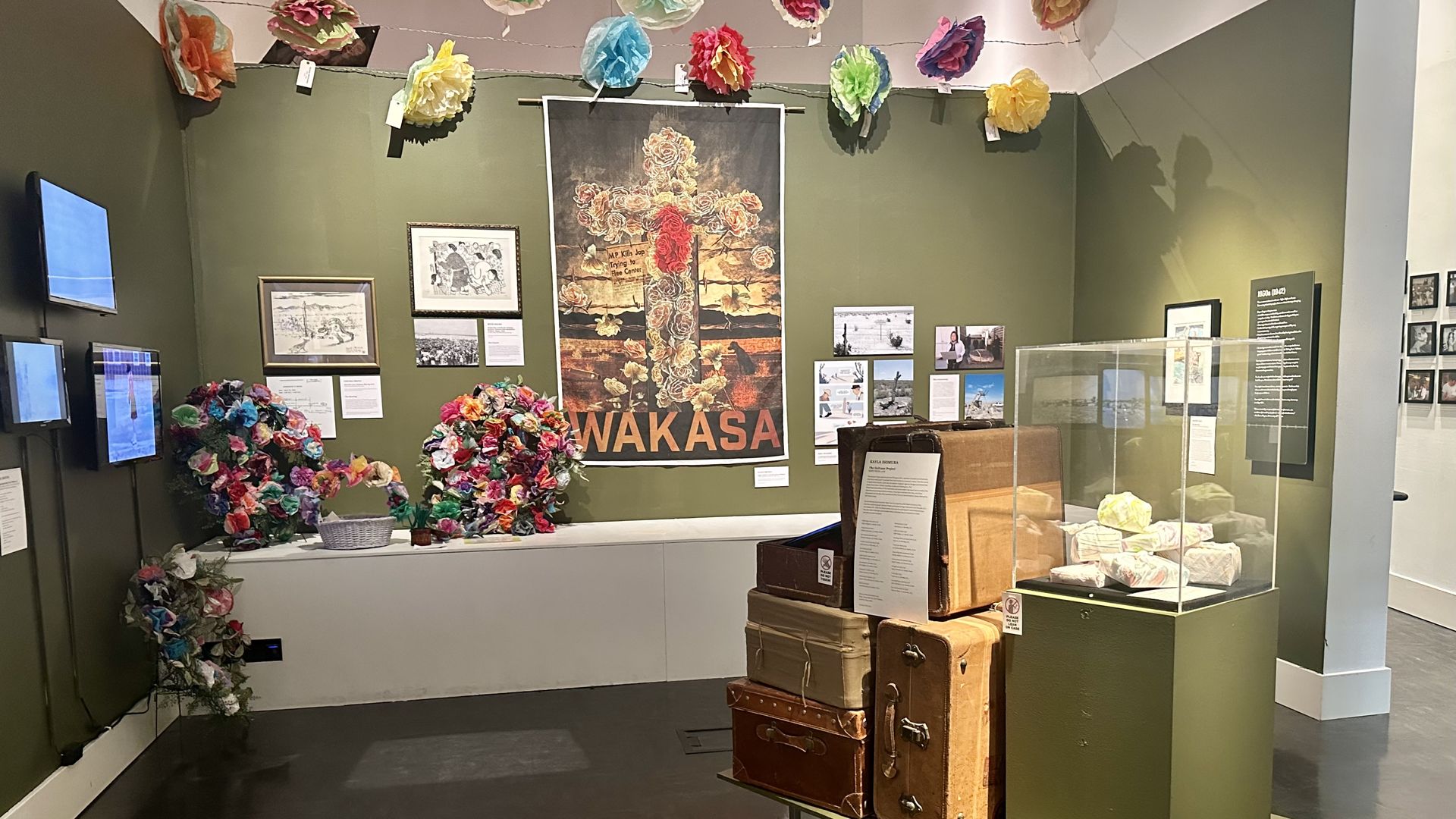 An image of a memorial on a back wall with paper flowers surrounding it, and a stack of suitcases on a display in a middle of a room. 