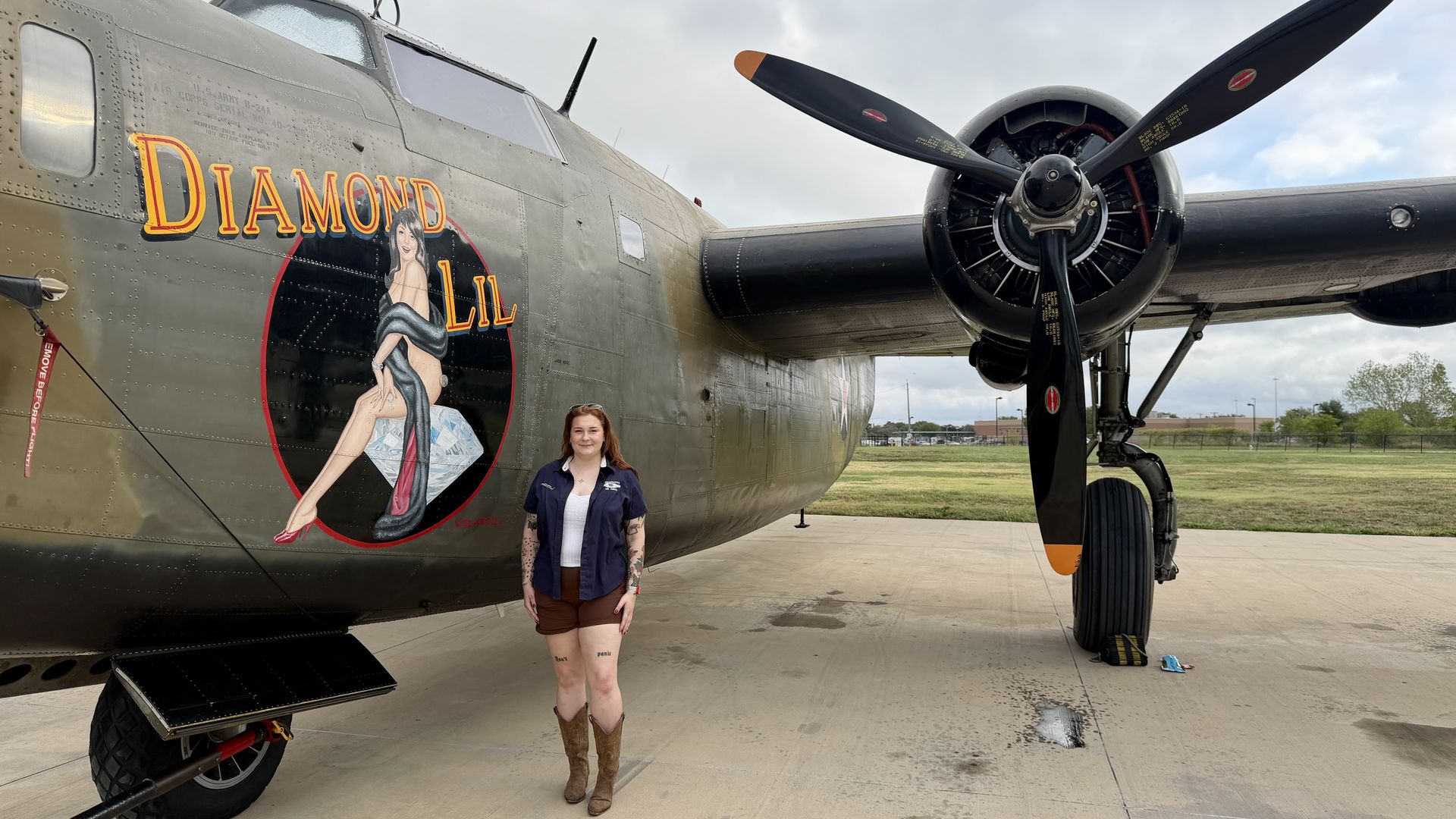 Side view of a vintage bomber named "DIAMOND LIL" with pinup art; a woman stands in front in a navy shirt and brown shorts; large black propeller engine on the right, airfield background.