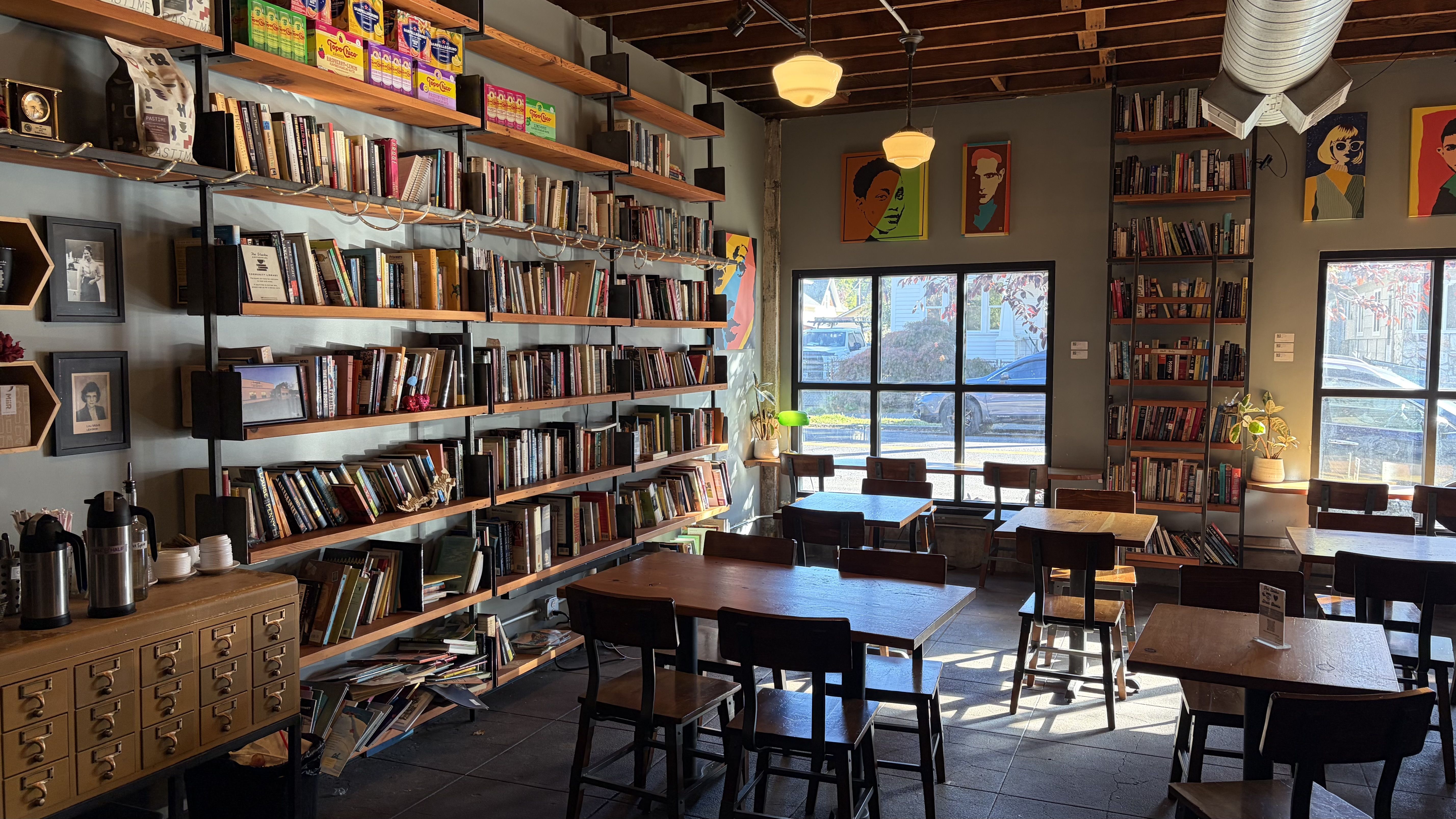Cozy bookstore cafe with long wooden bookshelves along the left wall, warm pendant lights, and ceiling beams. Sunlit windows on the right, tables and chairs, colorful posters, and potted plants.