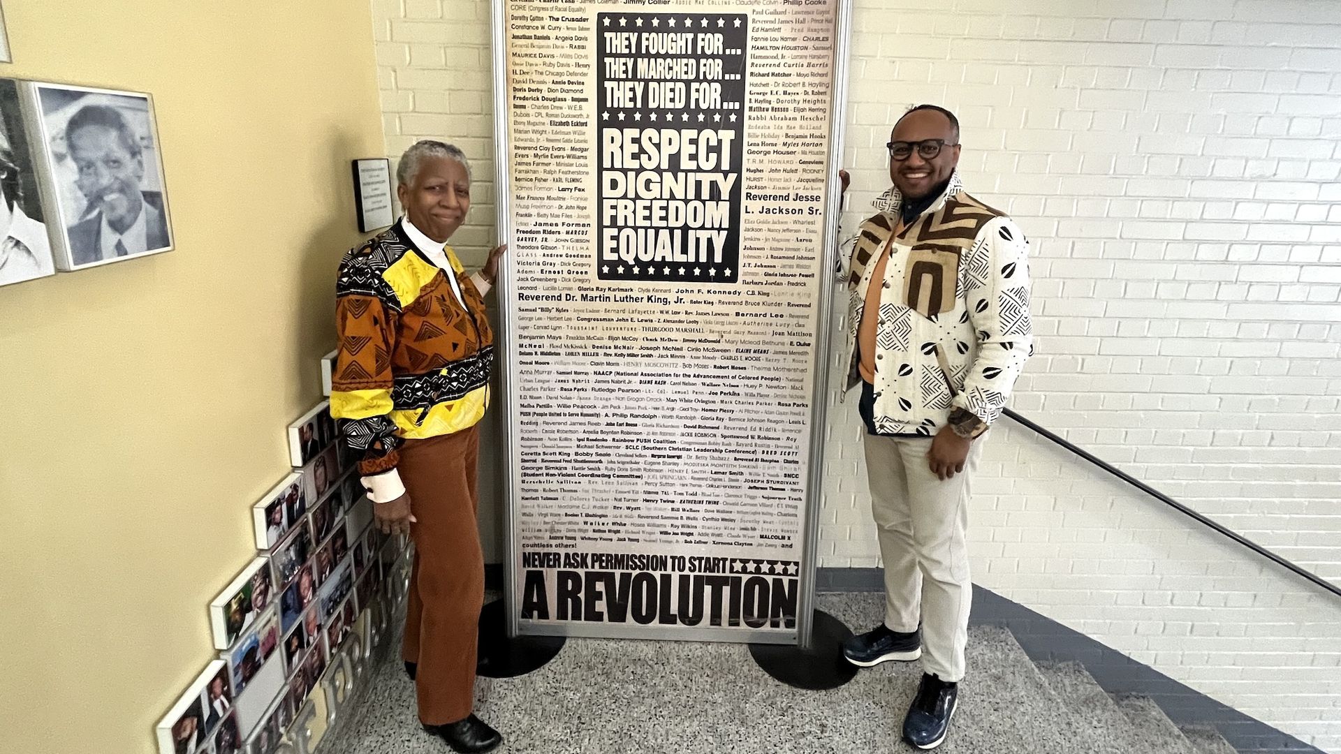 Two people stand beside a large poster titled "The Civil Rights Movement" with words like "Respect," "Dignity," "Freedom," and "Equality." The poster lists many civil rights leaders.