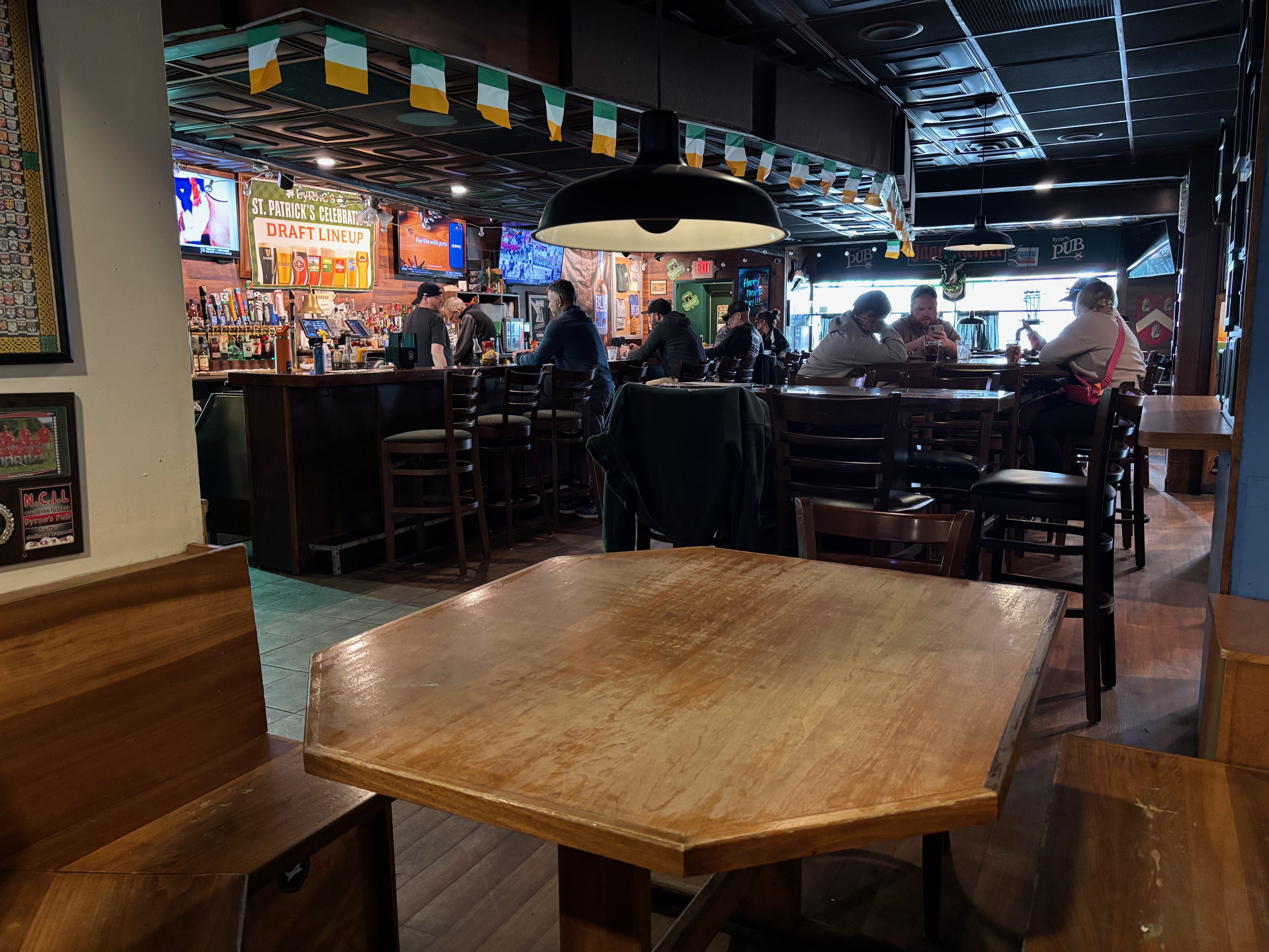 Pub interior with a hexagonal wooden table in the foreground, a long bar with stools, and patrons chatting; green-yellow bunting overhead and TVs behind the bar.