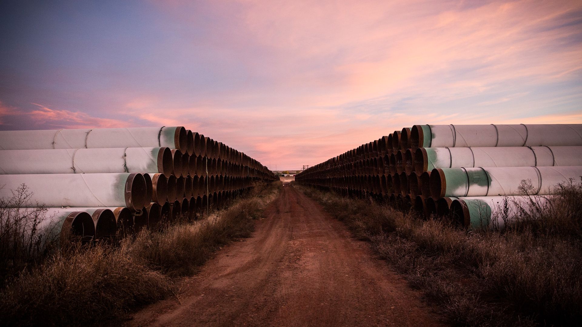 In this image, large pipes are stacked on either side of a dirt road.