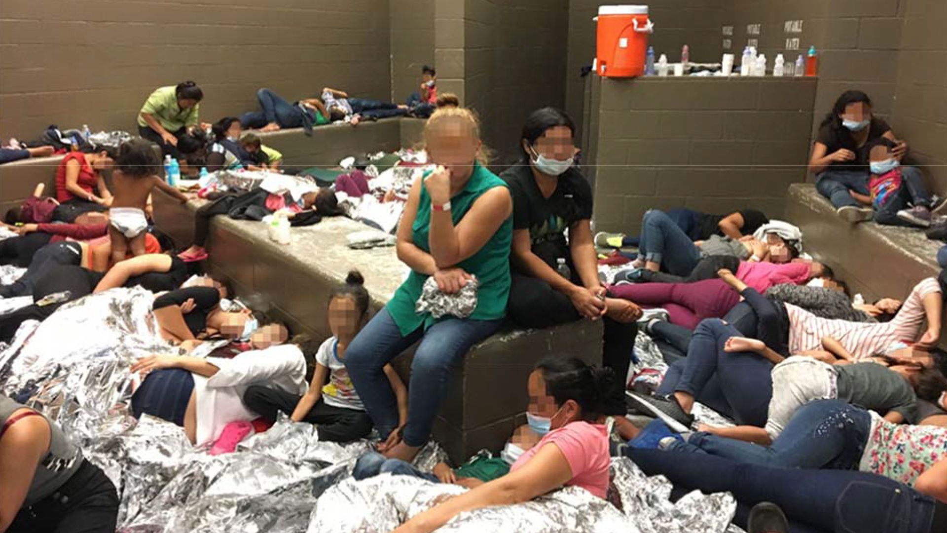 Women and child migrants crammed into a detention cell with aluminum blankets and blurred out faces. 
