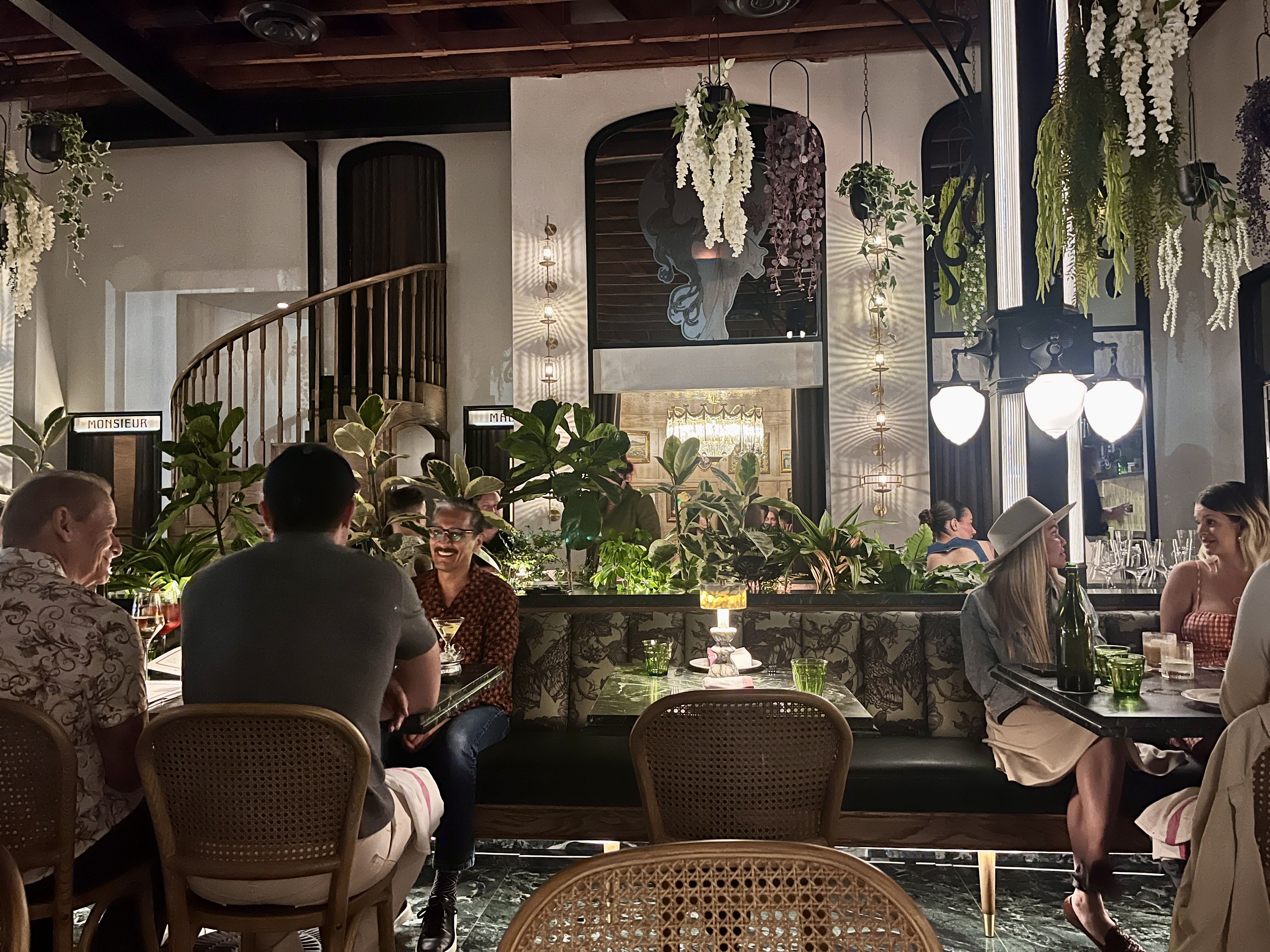 Cozy restaurant interior with a curved wooden staircase, hanging white flowers, and lush greenery. Diners sit at tables chatting over drinks under warm, ambient lighting.