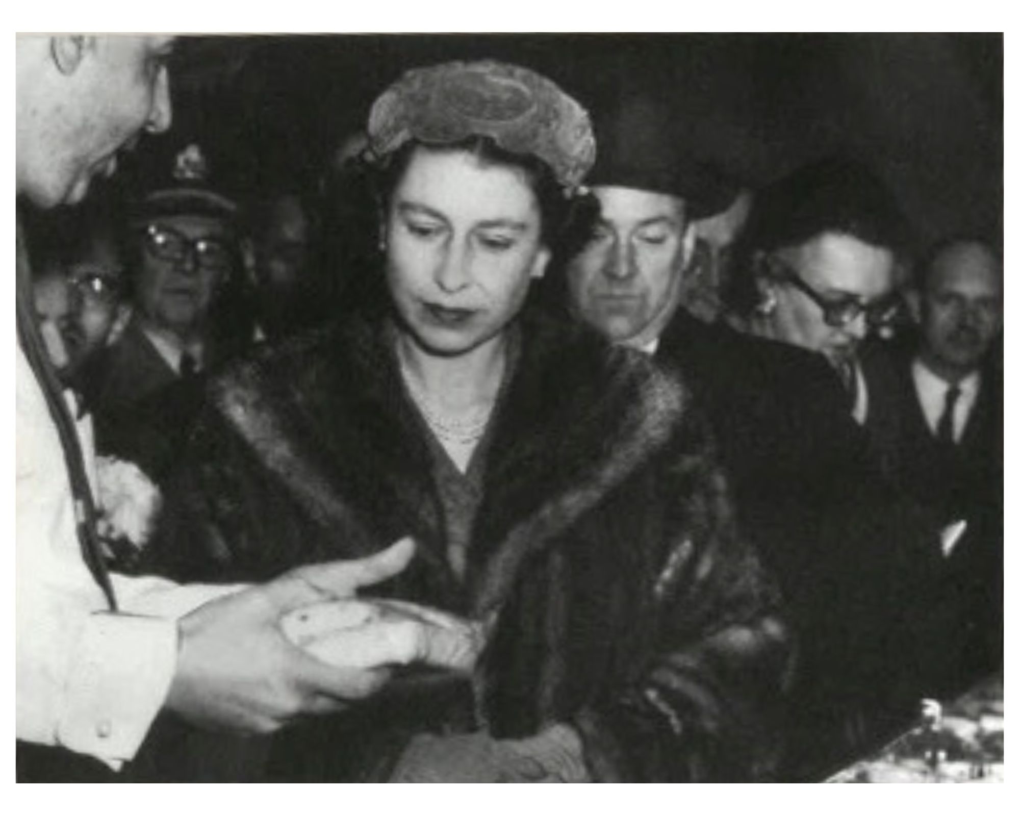 Black and white photo of Queen Elizabeth in a fur coat and hat looking at a dish held by a man in a white jacket, with several people in formal attire in the background.