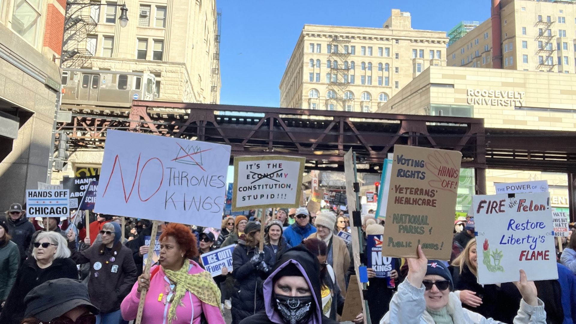 Crowd of protesters on a city street with signs reading "NO THRONES KINGS" and "HANDS OFF CHICAGO", an elevated railroad overpass, tall buildings, and Roosevelt University in view under a bright blue sky.