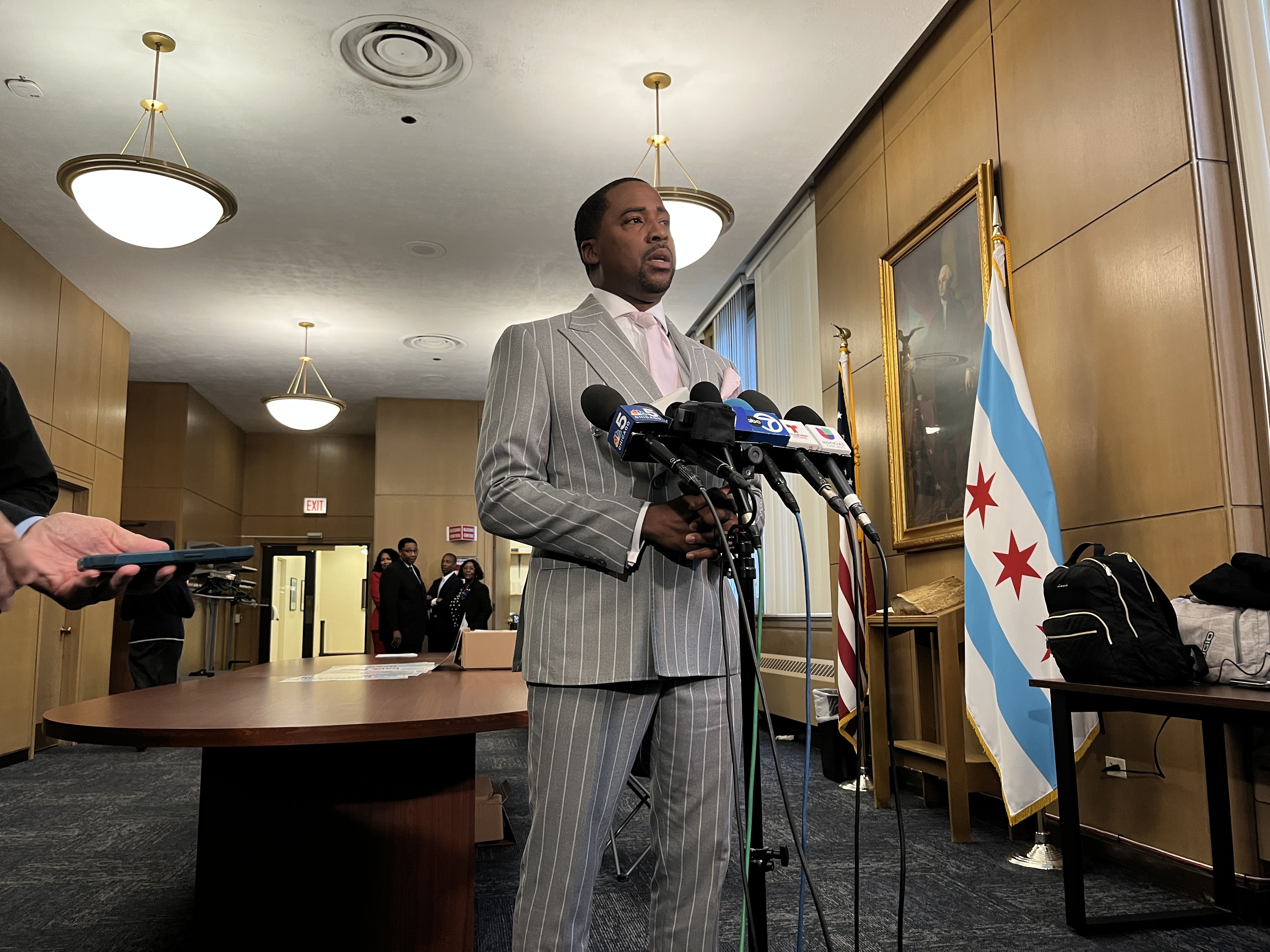 Man in a gray pinstripe suit and light pink tie speaking at multiple microphones in a wood-paneled room with Chicago and U.S. flags and a portrait on the wall.