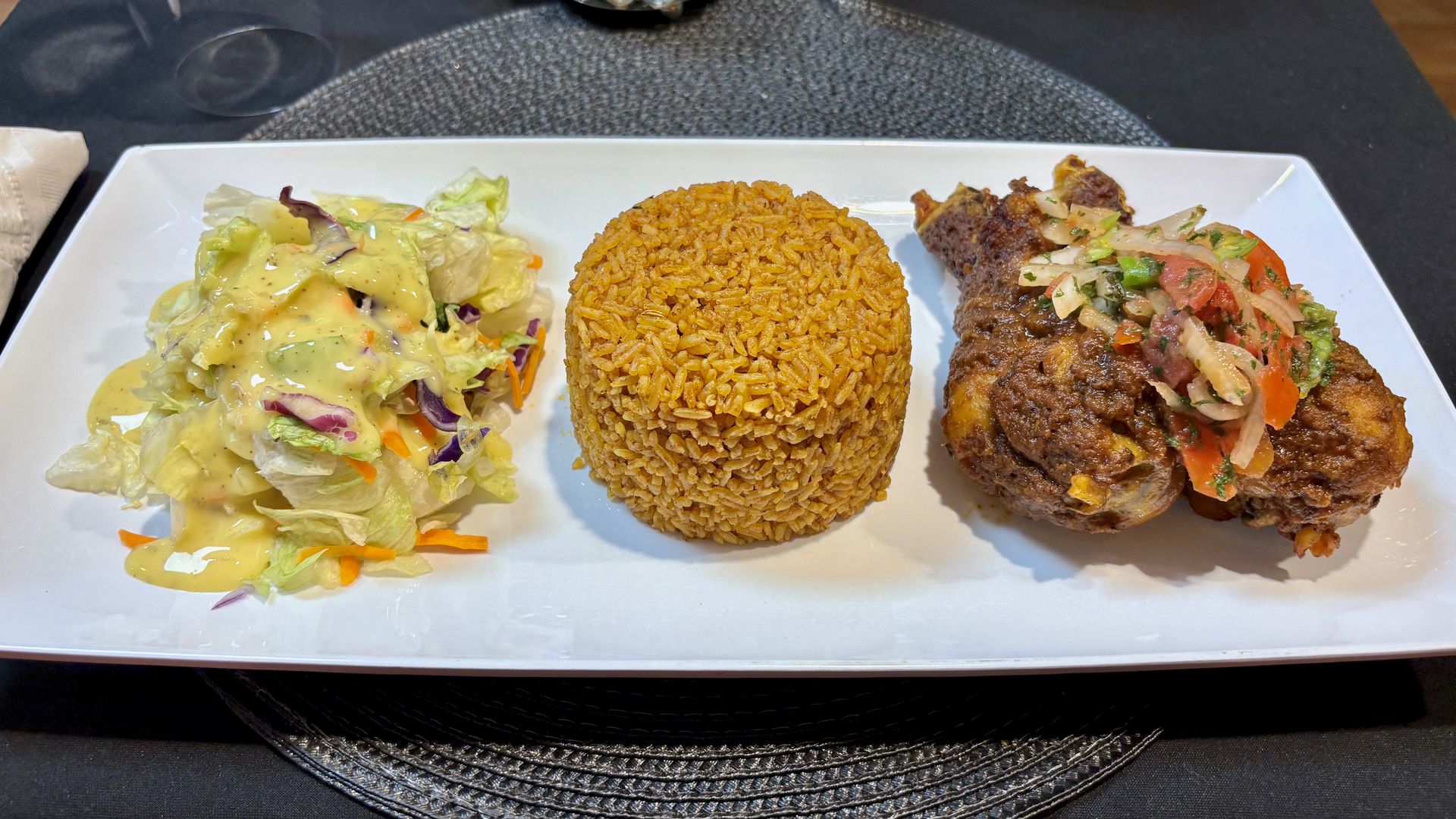 Rectangular white plate with a scoop of seasoned jollof rice, two pieces of baked chicken topped with peppers and onions, and a side salad with creamy yellow dressing.