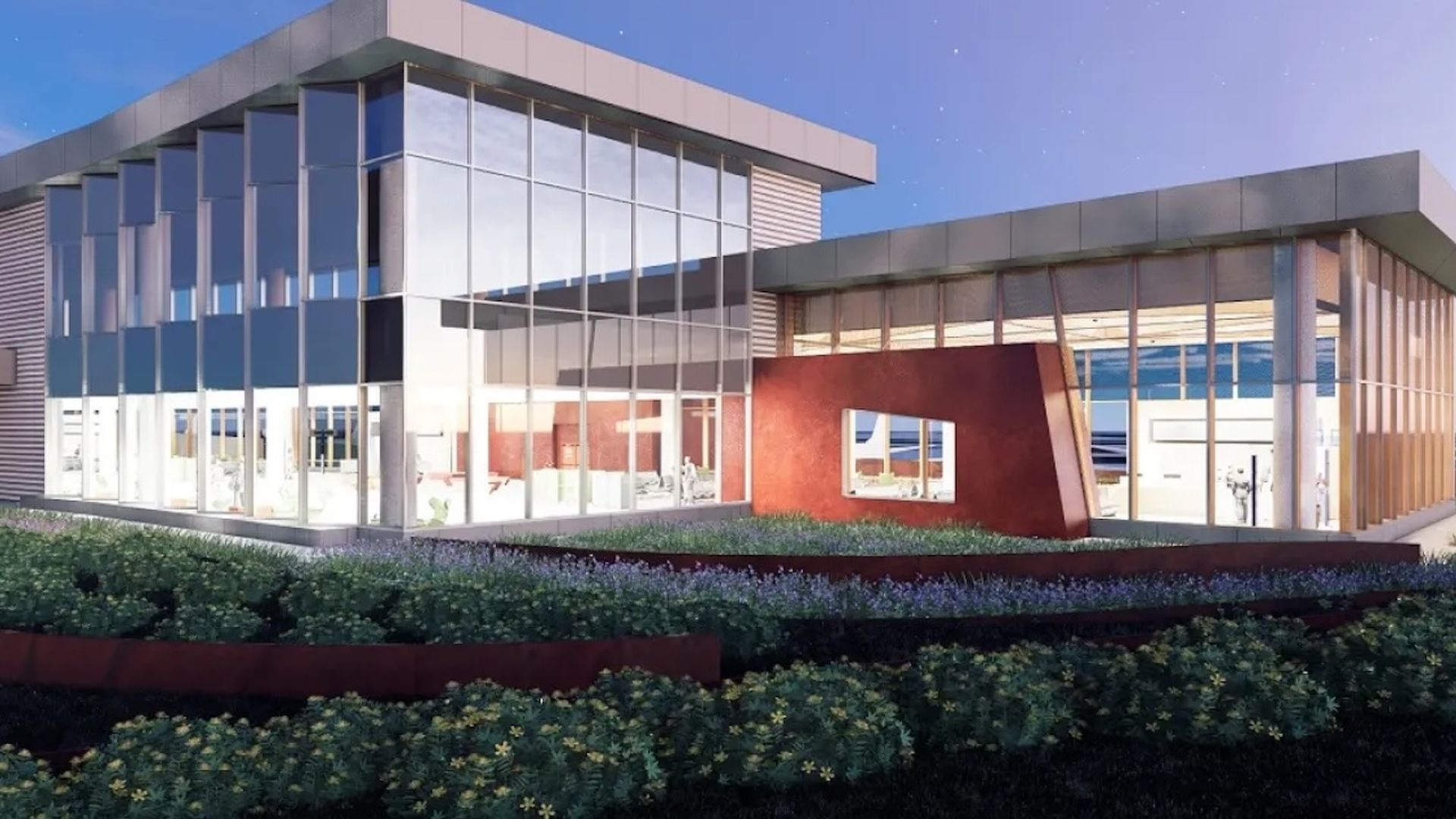 Modern building with large glass windows, a rust-colored wall feature, green shrubs, and purple flowers under a clear blue sky at dusk.