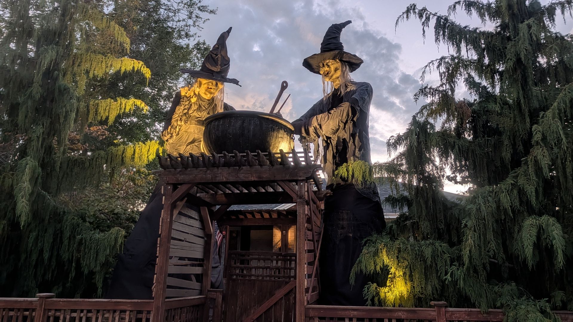 Two large witch statues in black robes stand beside a cauldron on a wooden platform, among tall evergreen trees at dusk; a small American flag is visible in the lower left.