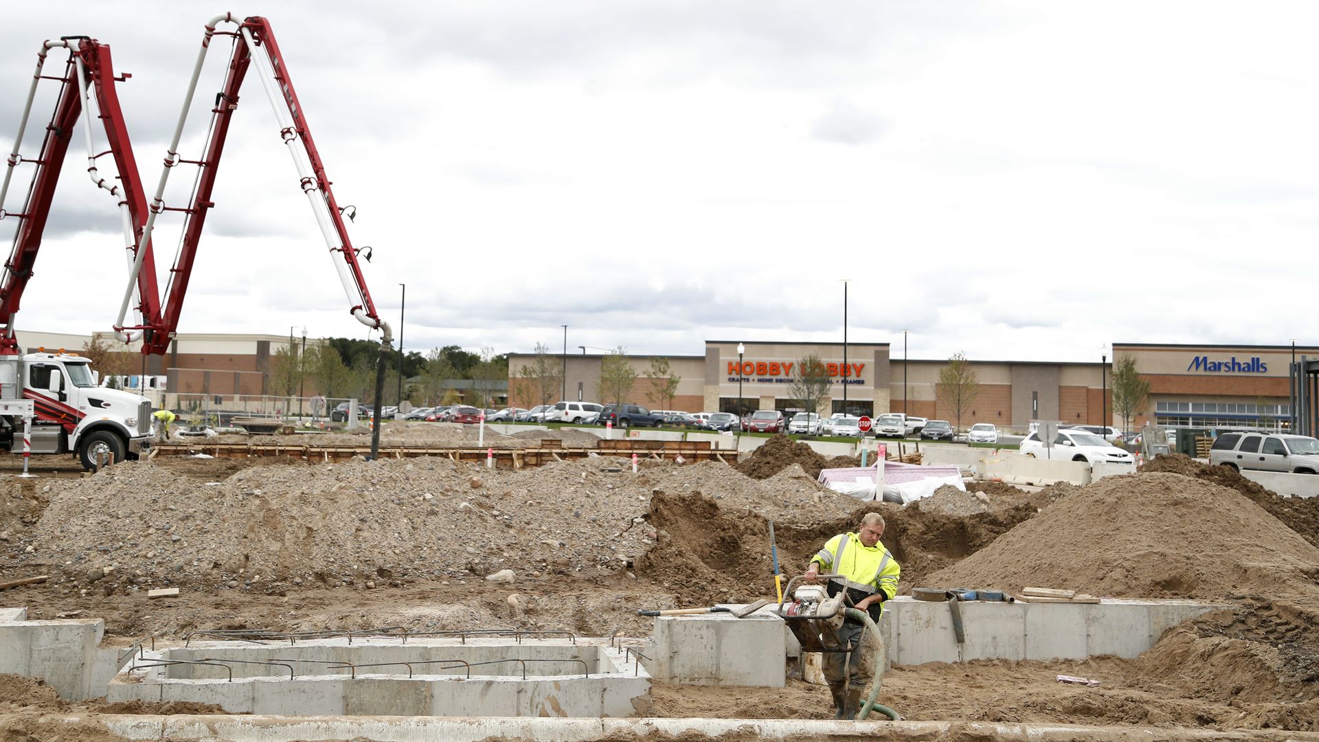 Construction site with a large red concrete pump truck on the left, dirt piles and concrete forms in the foreground, Hobby Lobby and Marshalls in the background; worker in a yellow vest stands nearby.