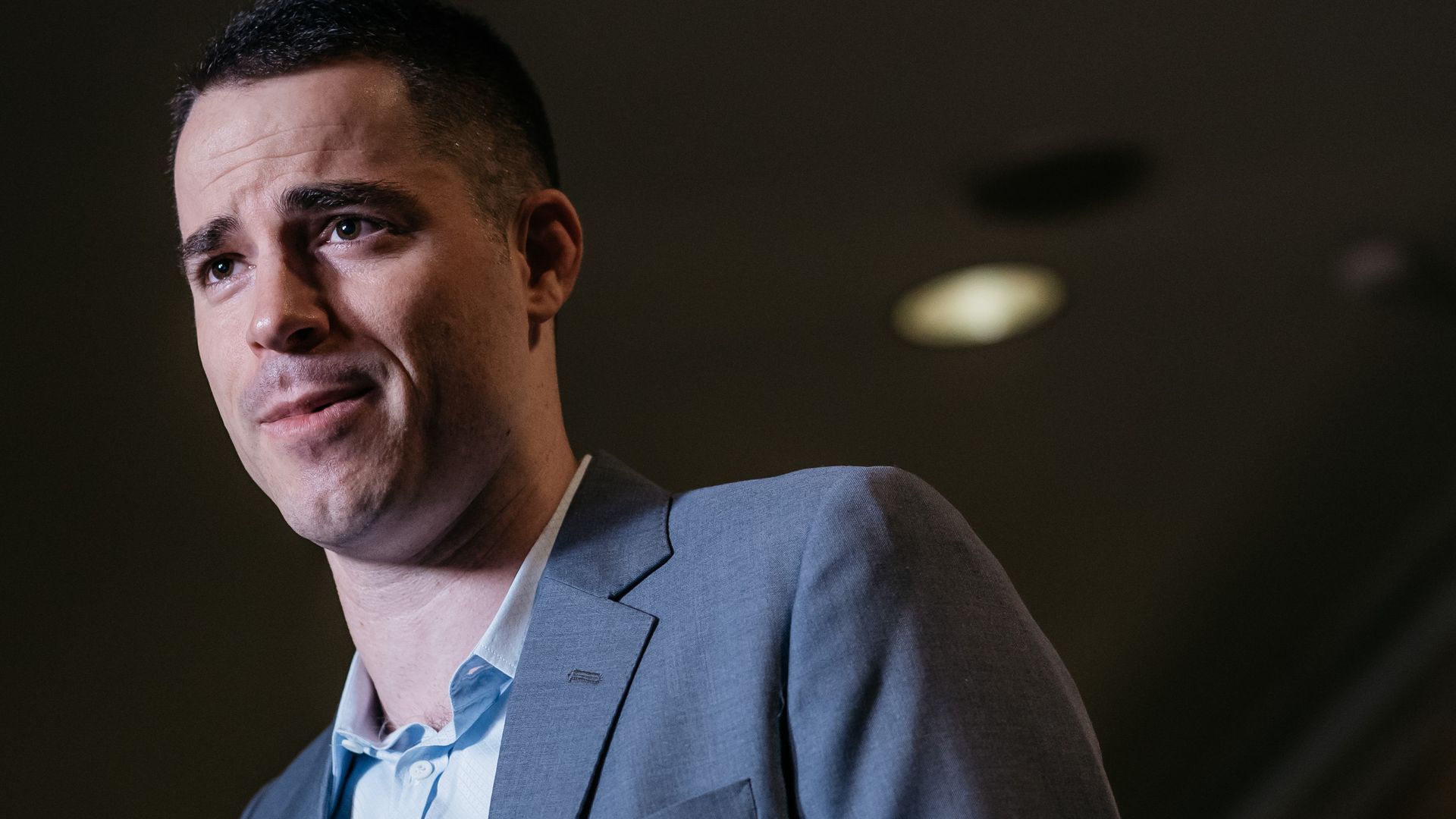 A man in a suit without a tie, and very short her, speaking at a conference. From shoulders up. Slightly turned to the side. 