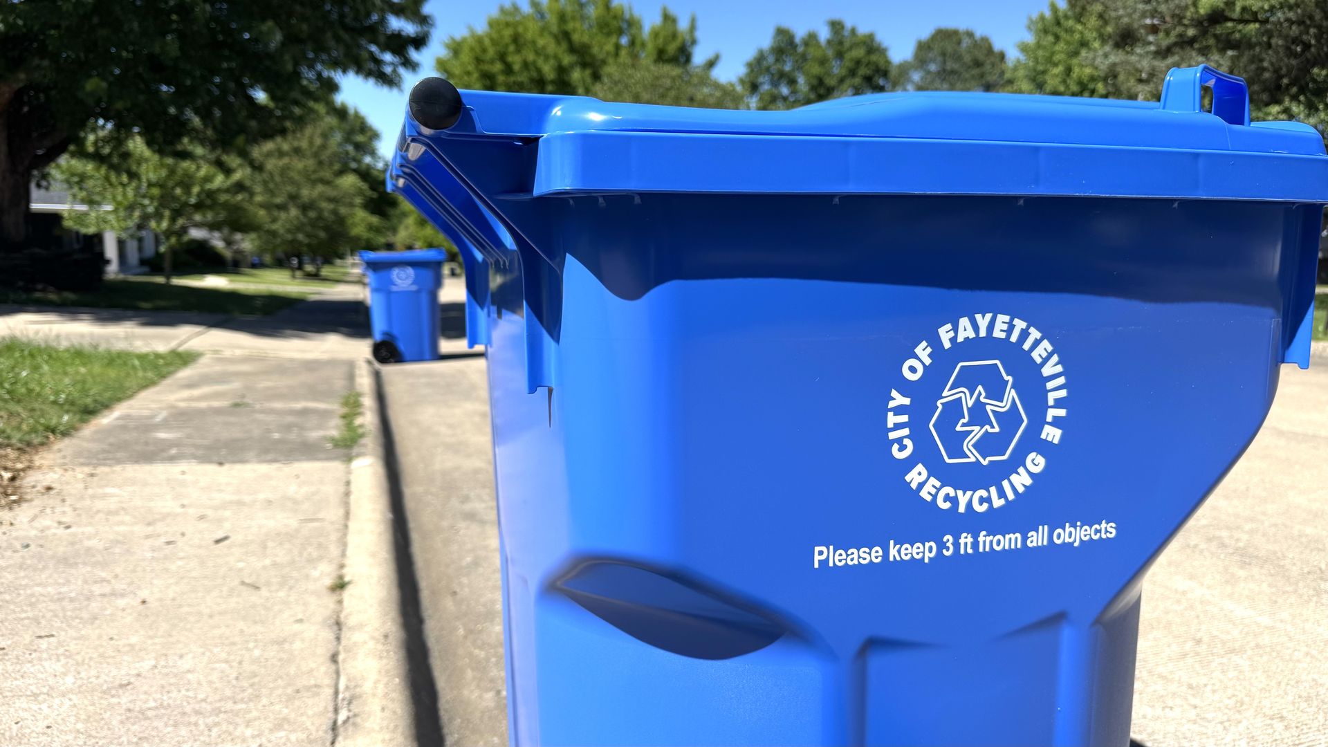 Blue City of Fayetteville recycling bins line a residential street on a sunny day.