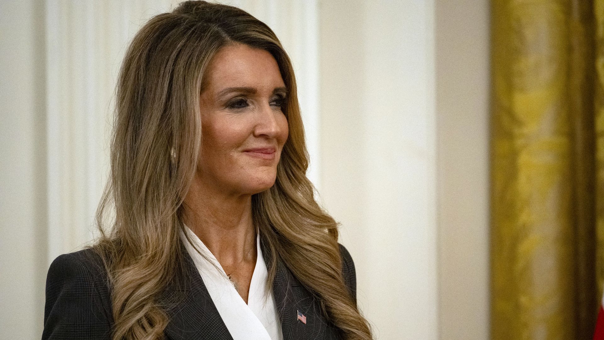 Kelly Loeffler smiles as she looks on while inside the White House during an event. She is wearing a dark blazer, a white blouse and an American flag pin, and her hair is down.