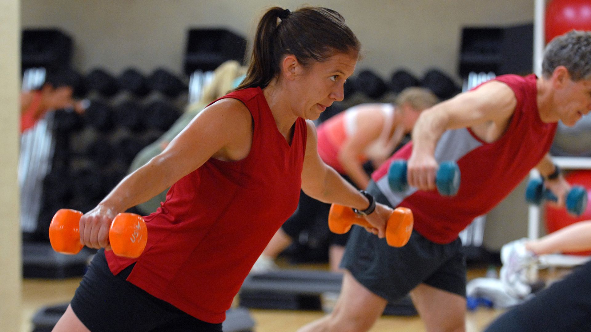 An exerciser balances on one foot with orange weights in each hand
