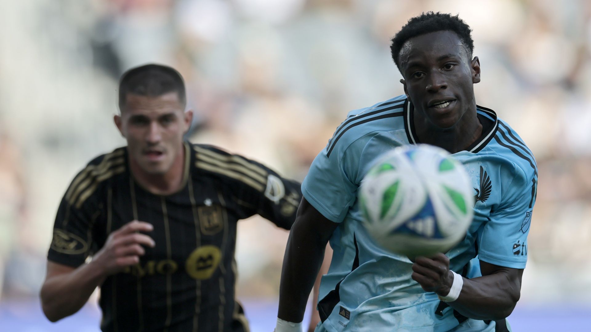 A soccer player in a light blue jersey charges after a ball that hovers in the foreground