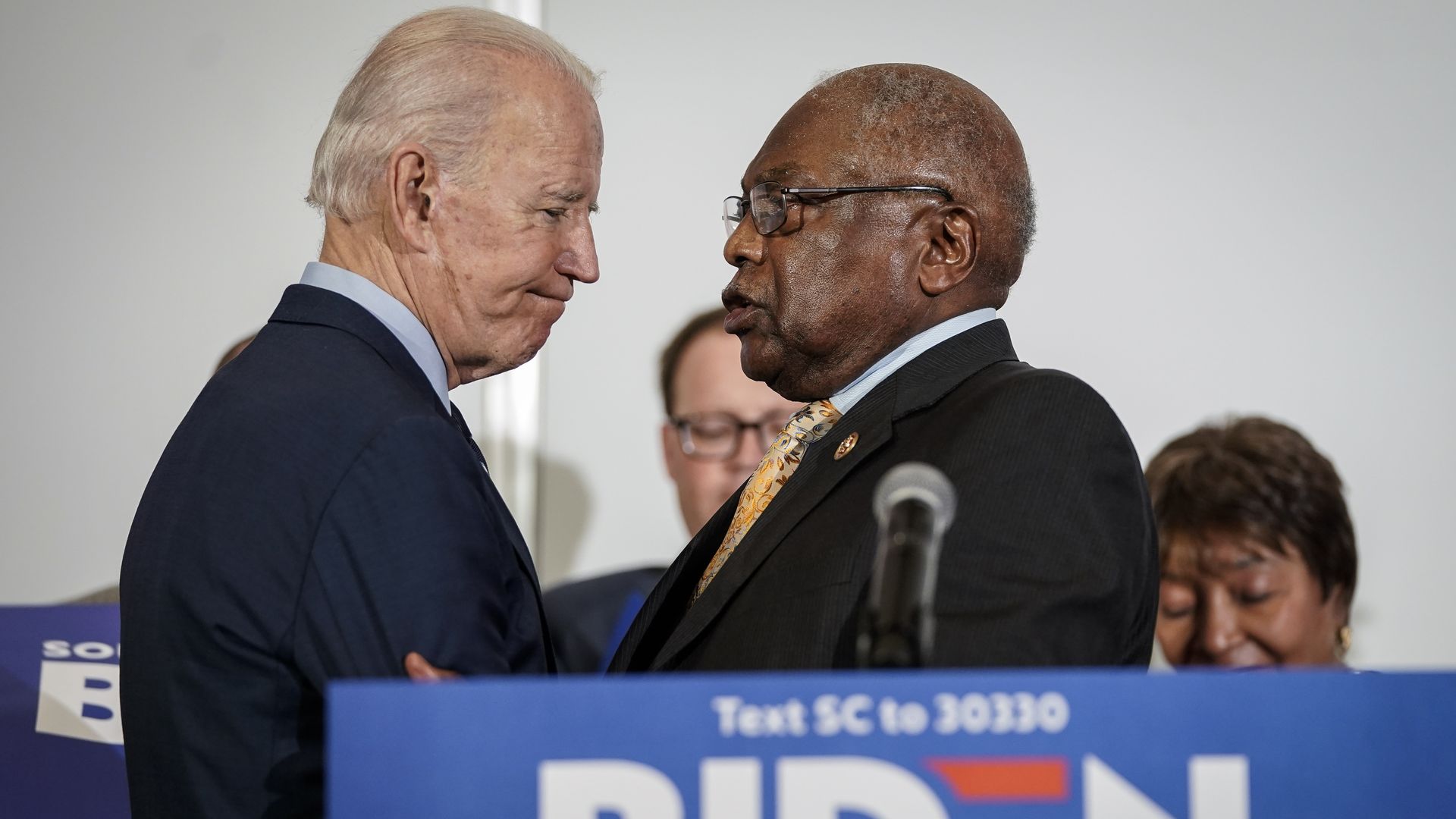 In this image, Biden and Clyburn hold each others' arms while standing in front of a campaign podium
