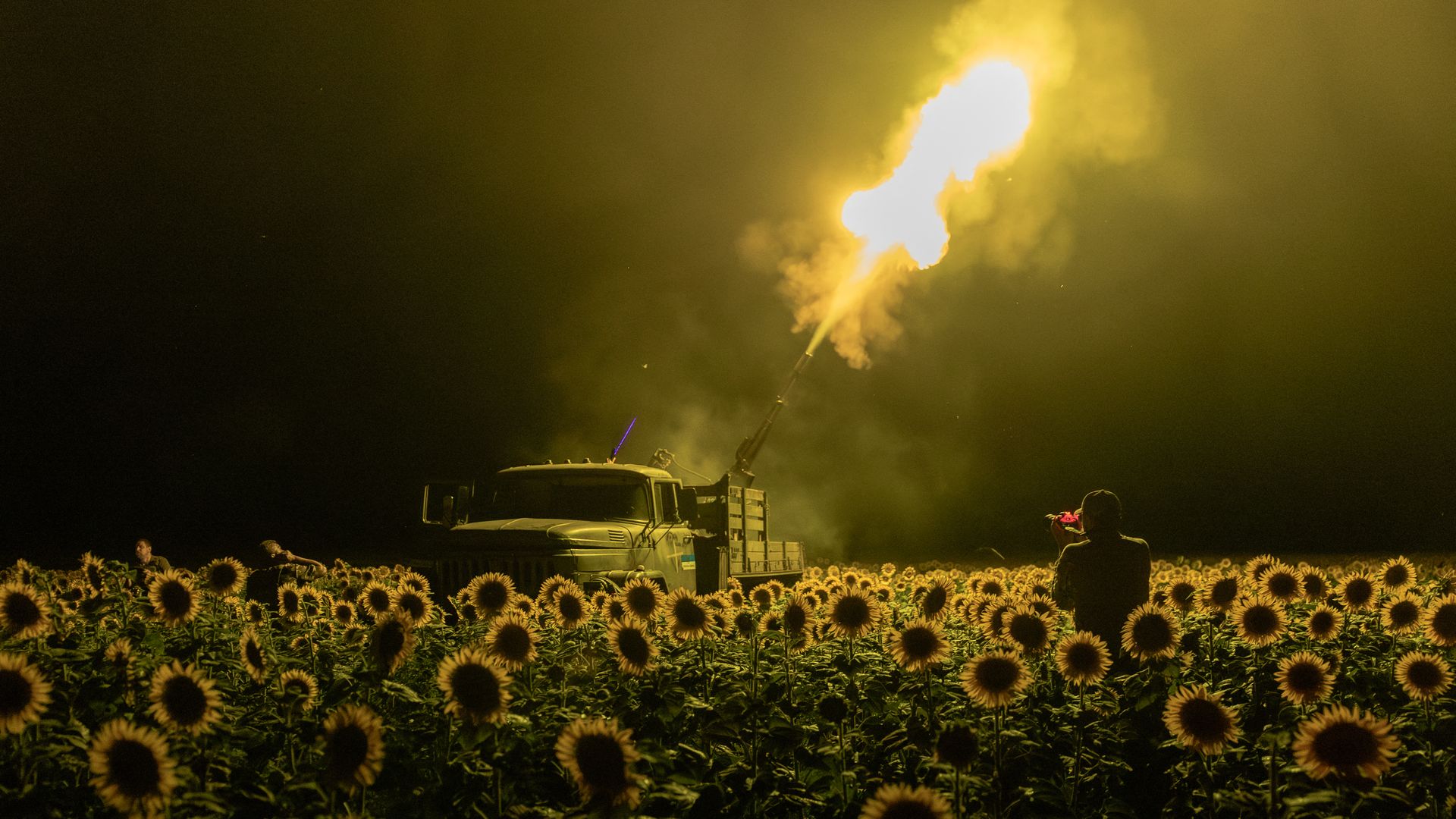 A military truck firing a large gun at night in a field filled with sunflowers, illuminated by the bright muzzle flash and surrounded by smoke and silhouettes of people.