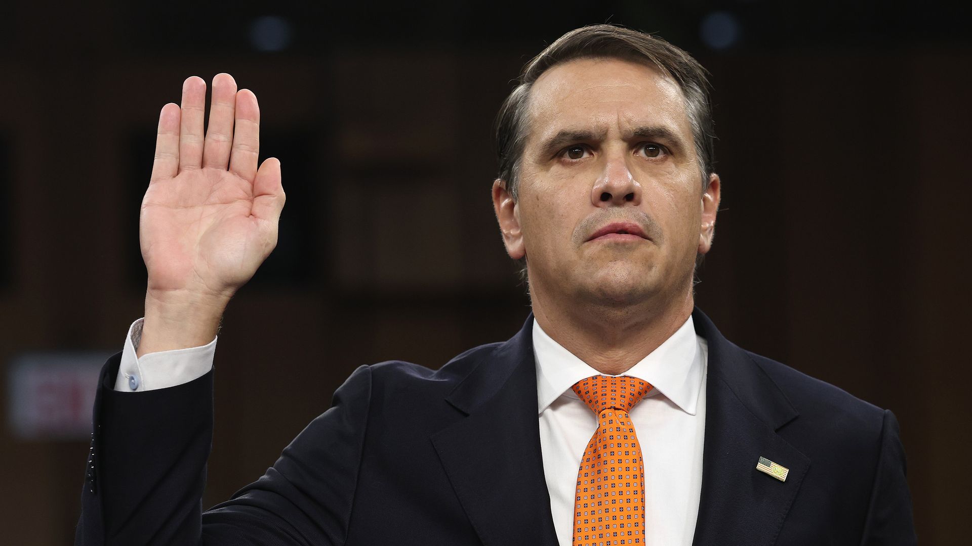 Todd Blanche, a man in a dark suit and orange tie with a small American flag lapel pin, raises his right hand in a ceremonial oath.
