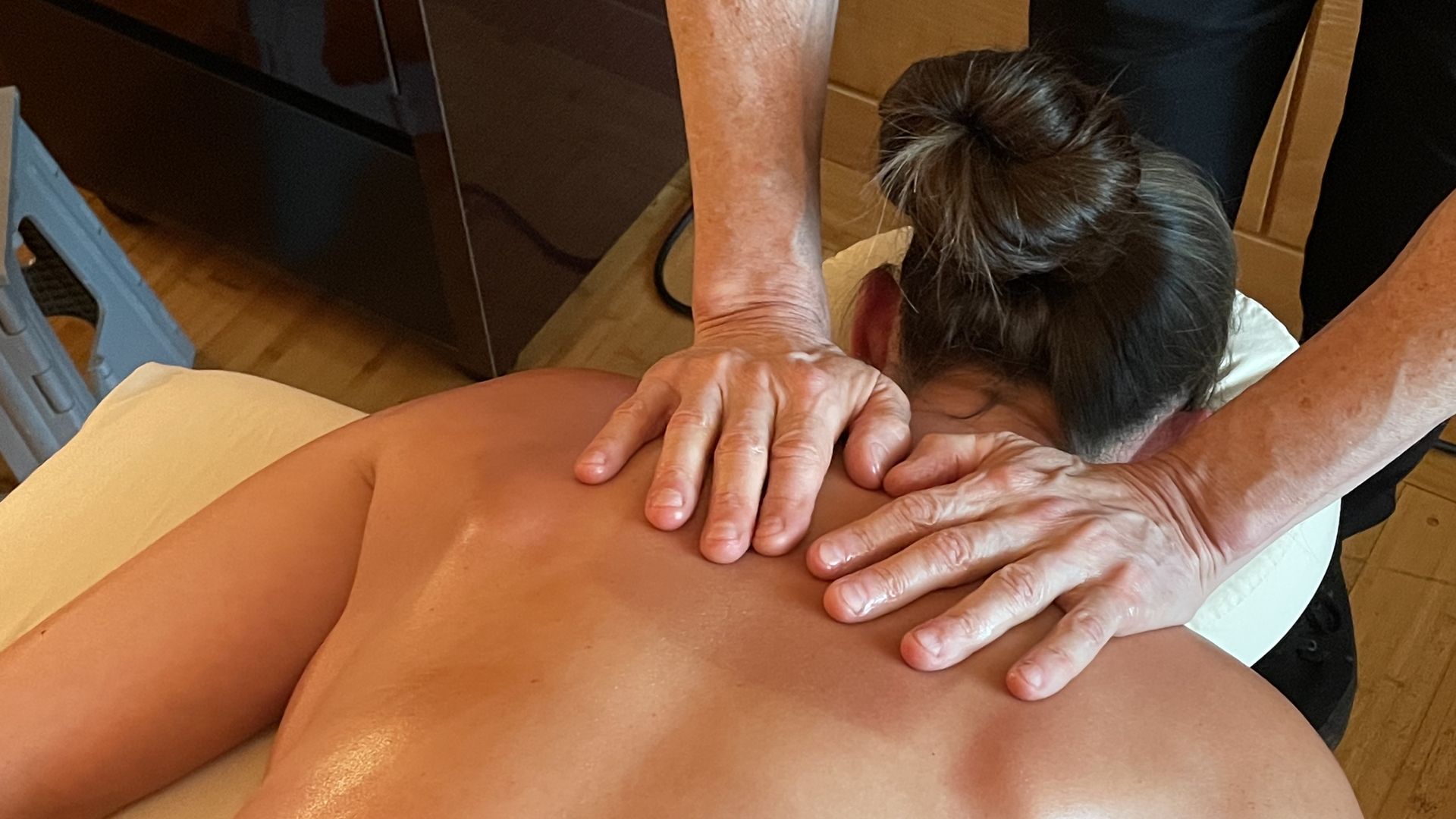 A massage therapists' hands rest on his client's bare shoulders as he prepares to rub her back with CBD massage oil.