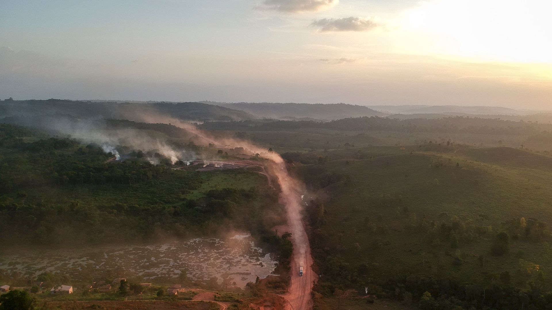 A picture taken from an airplane of a road through the Amazon, pockmarked by smoke in the distance.