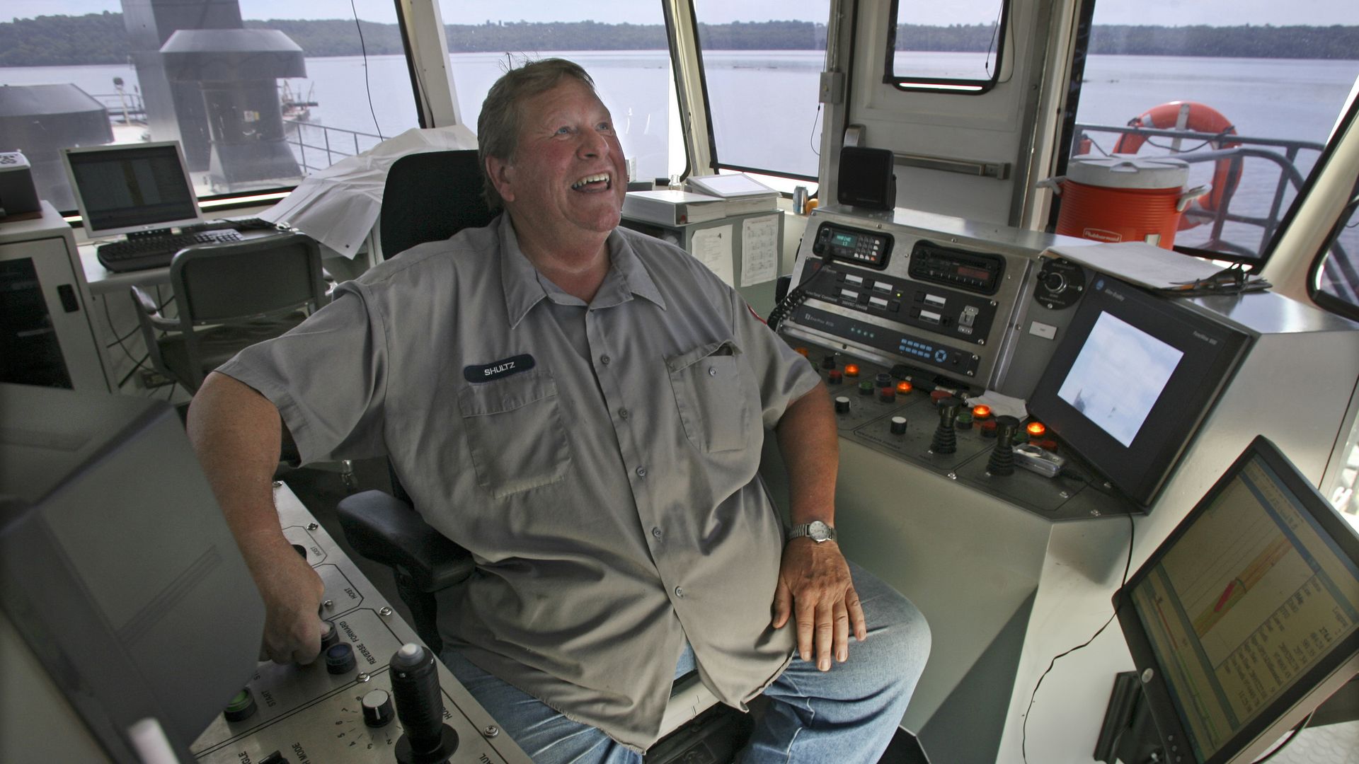 Smiling man in gray shirt and jeans seated in a ship's control room surrounded by monitors, controls, and windows showing a calm river and distant shoreline.