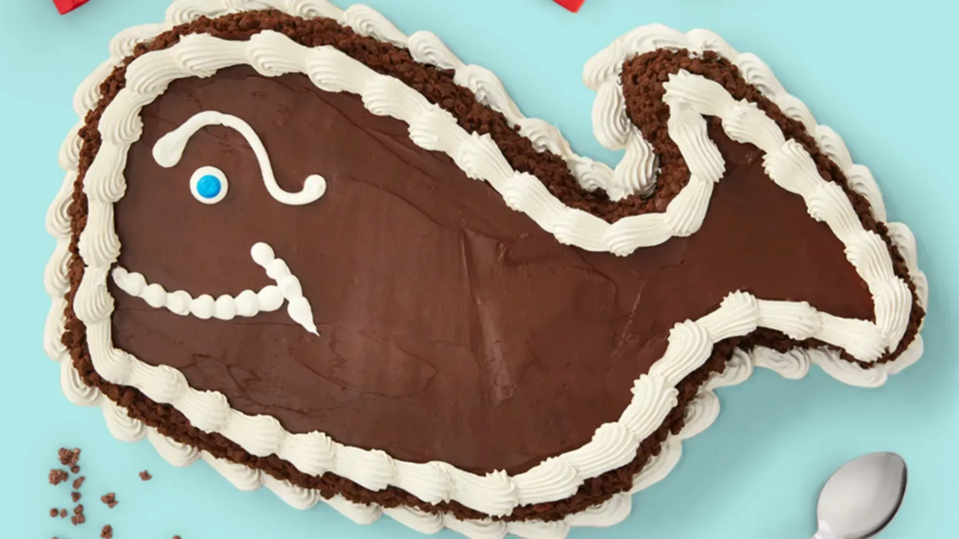 Close-up of a dinosaur-shaped chocolate cake on a teal background, with white frosting outlining the creature and a blue eye; chocolate crumbs around the edge, and a spoon nearby.