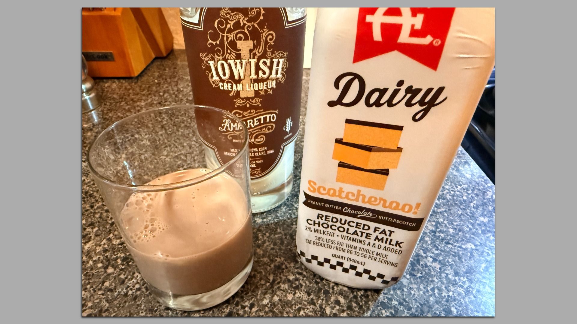Frothy chocolate drink in a glass sits on a speckled kitchen counter, flanked by a brown ornate liqueur bottle and a white 'Dairy' carton with orange stripes in the background.