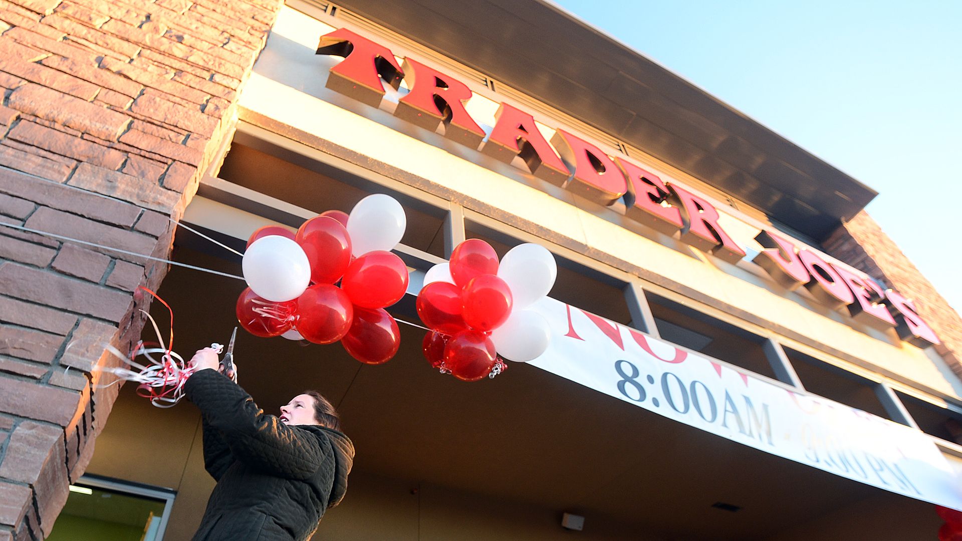 Person in a dark jacket ties red and white balloons to a storefront during a grand opening; brick pillar on the left, large red letters above, banner reads "8:00 AM".