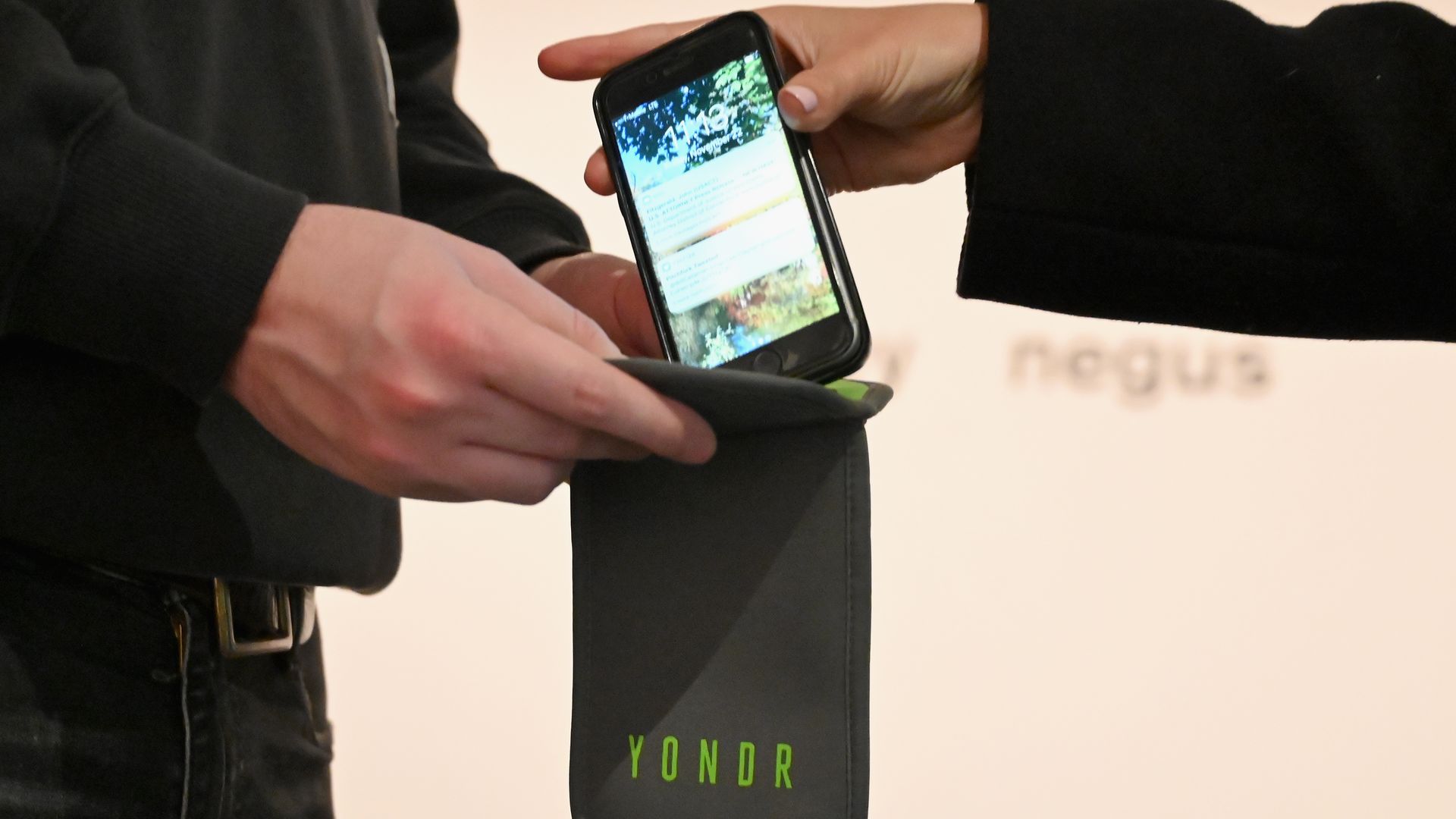 Two hands insert a smartphone into a black YONDR sleeve; the screen shows apps while a green YONDR logo is visible on the case.