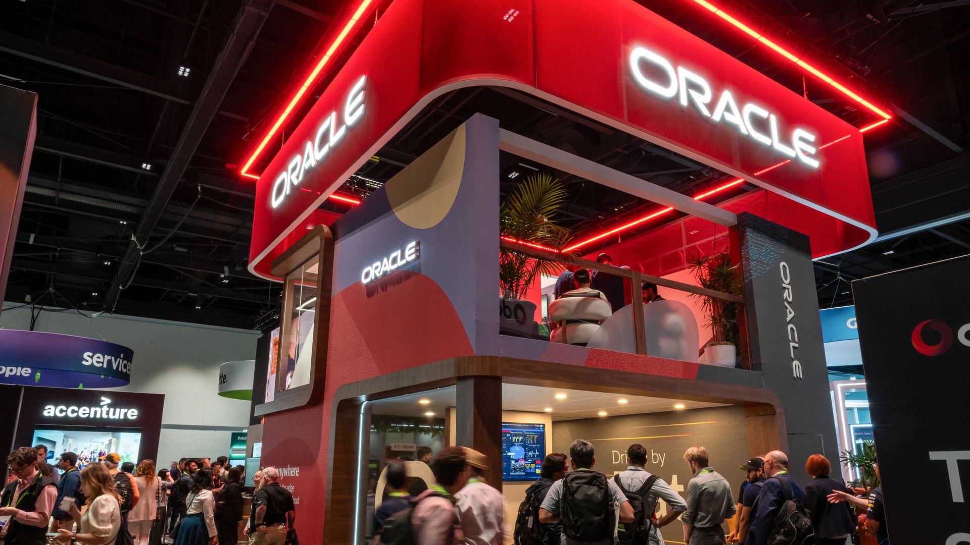 Oracle booth at a tech expo: a two-level, red neon-framed display with large ORACLE signs; visitors crowd the floor, viewing screens and an upstairs seating area with plants.
