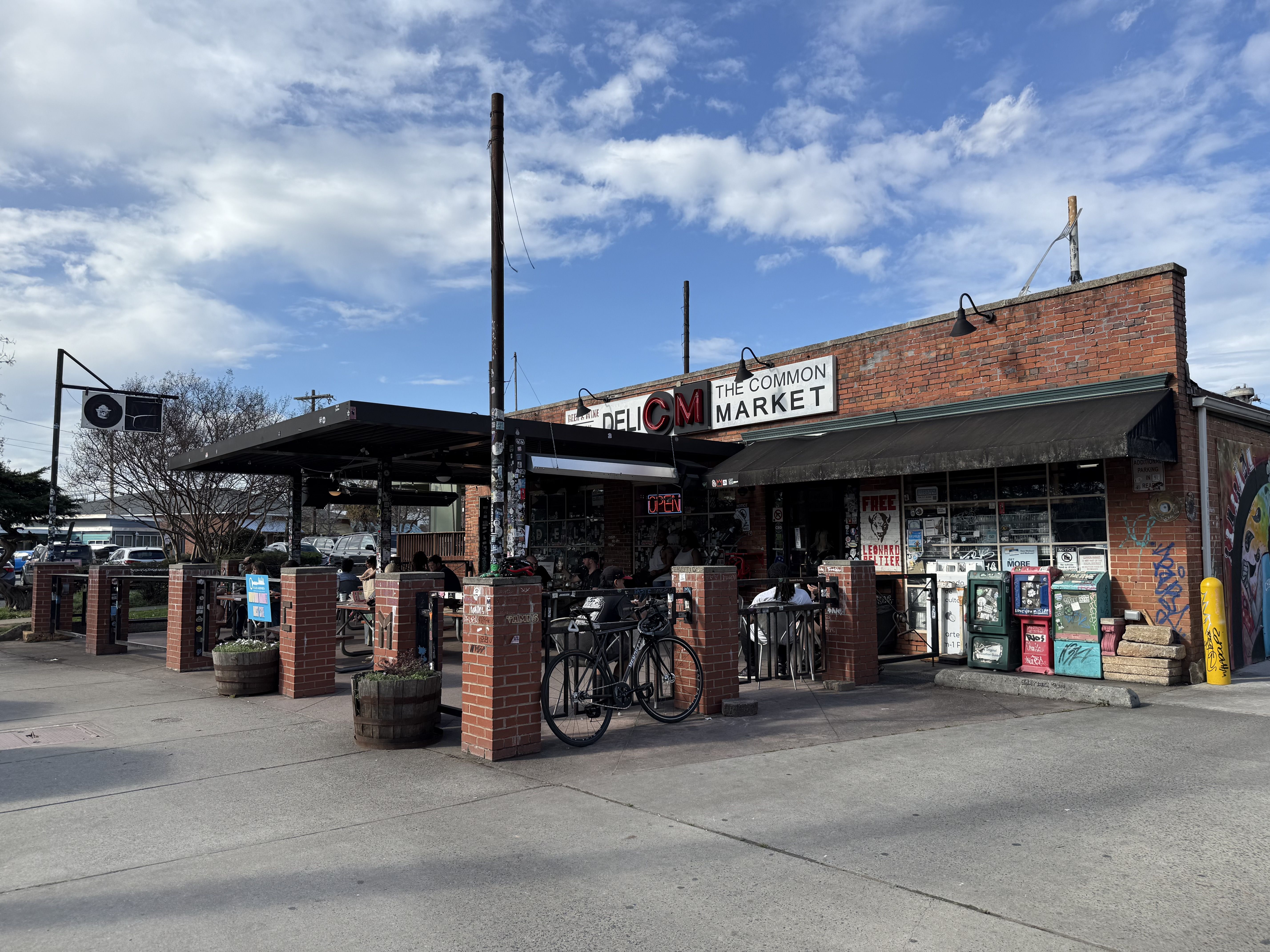 Brick storefront with sign \"The Common Market\" and a black awning; outdoor seating under a canopy, people dining. Bicycles lean on brick pillars; graffiti wall and vending machines on the right, blue sky above.