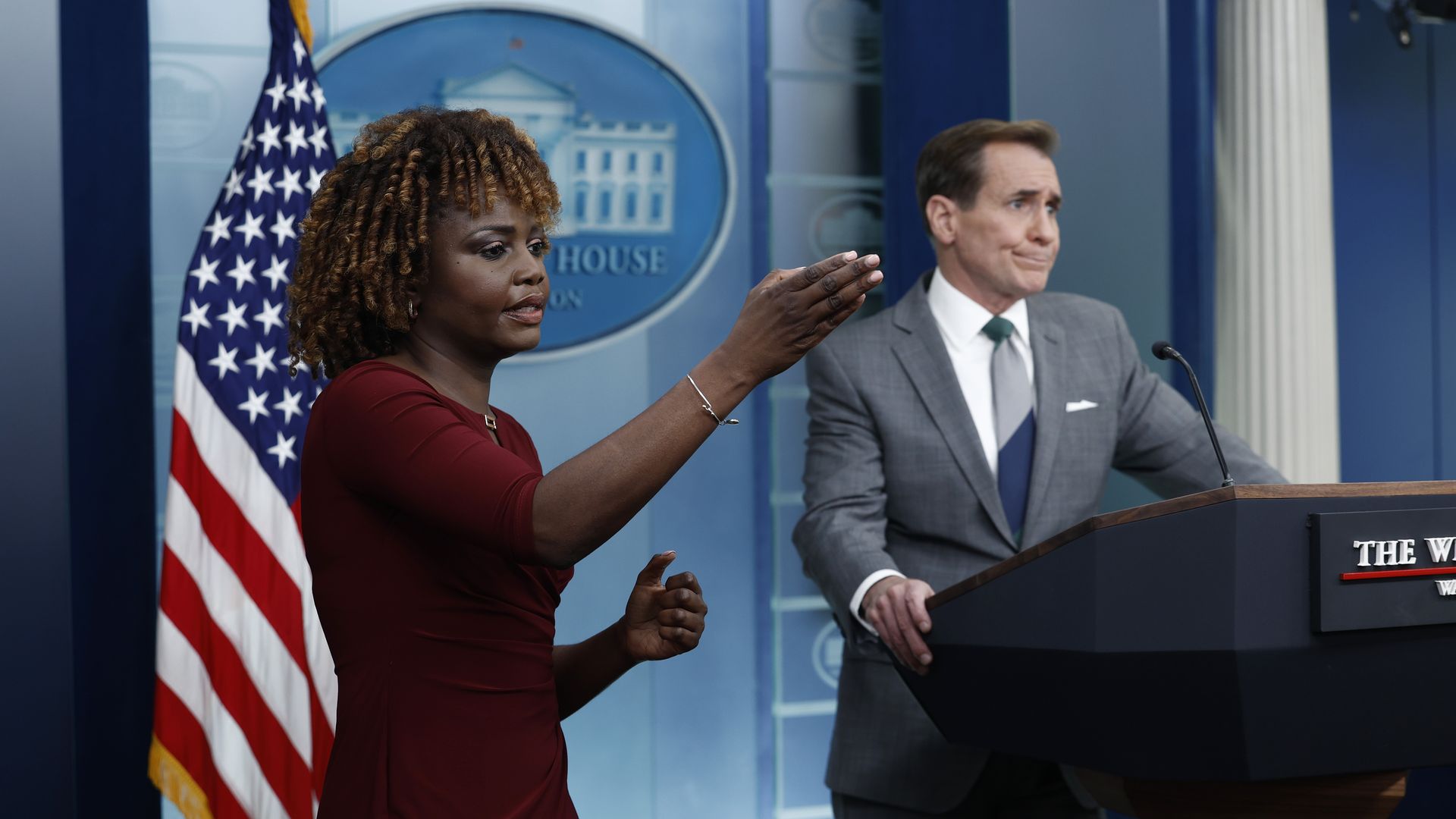 Karine Jean Pierre, wearing a red dress, and John Kirby, wearing a gray suit, in the White House briefing room.