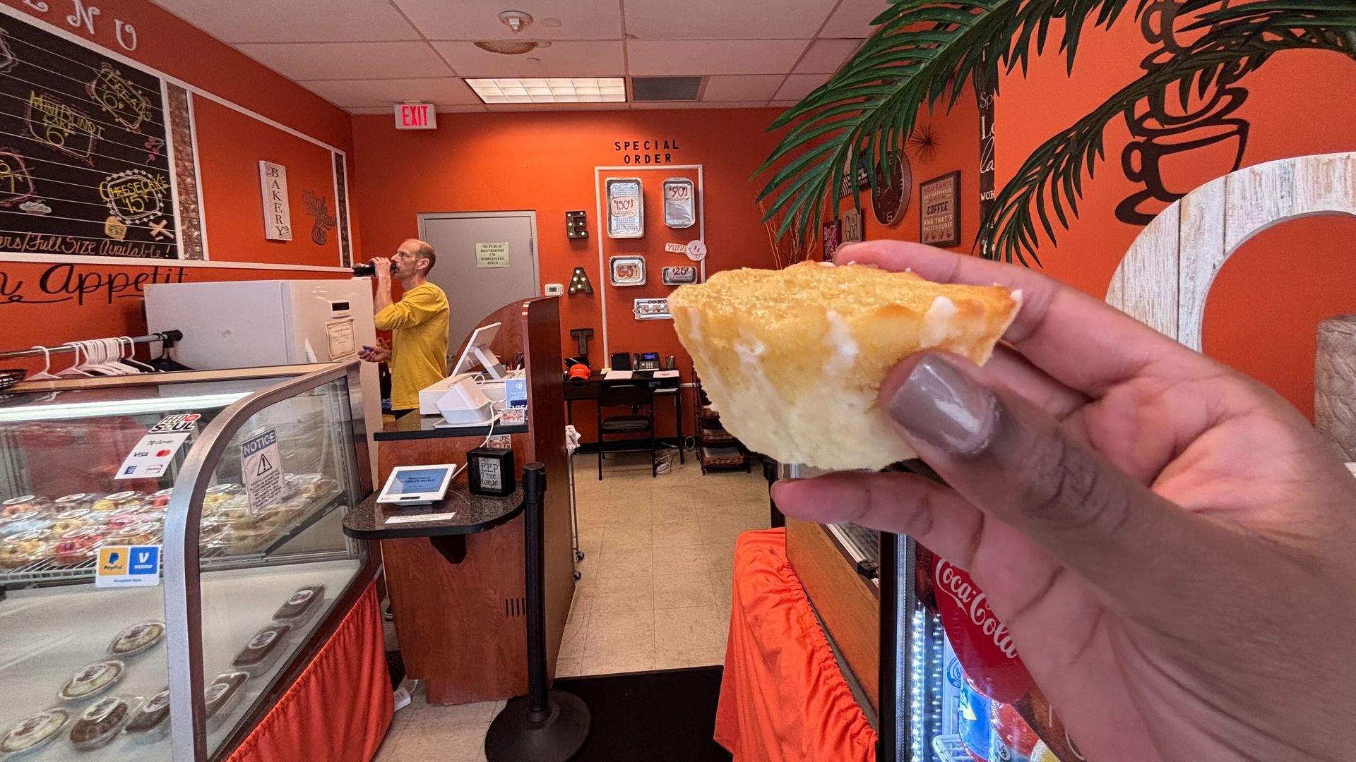 Inside a bright orange bakery, a foreground hand holds a crusty bread slice; in the back, a man in a yellow shirt drinks from a bottle near the counter, with a pastry display and a palm mural.