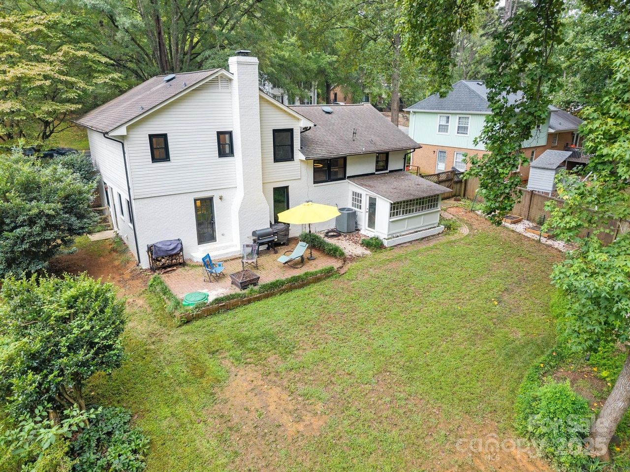 Backyard of a two-story white house with black window frames, a small patio with chairs, a yellow umbrella, a grill, and a grassy yard surrounded by trees and neighboring houses.