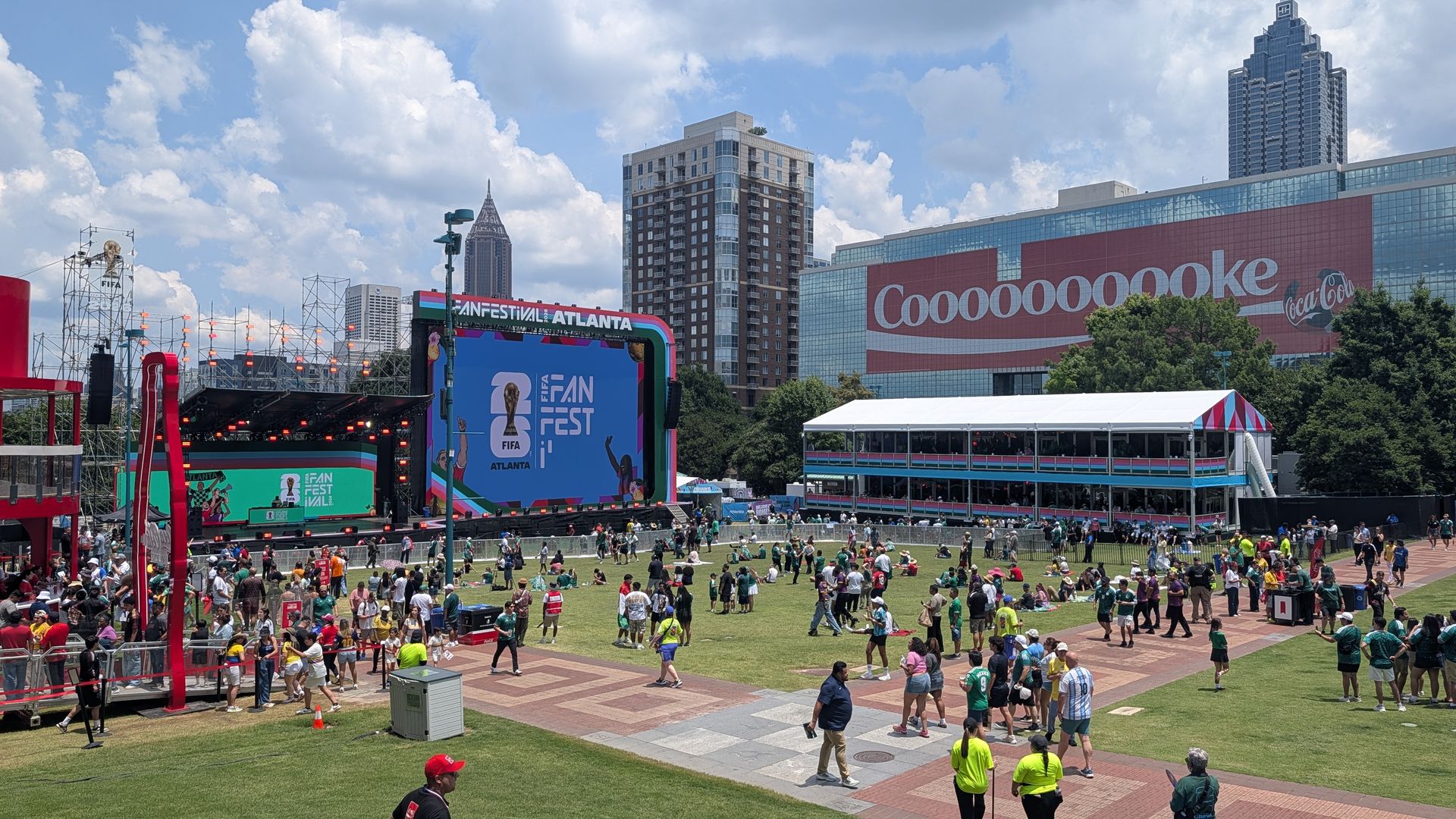 Outdoor festival in Atlanta with a large stage and colorful screens showing "FIFA Fan Fest Atlanta"; a red Coca-Cola mural on a glass building to the right; crowds on a grassy lawn under a blue sky.