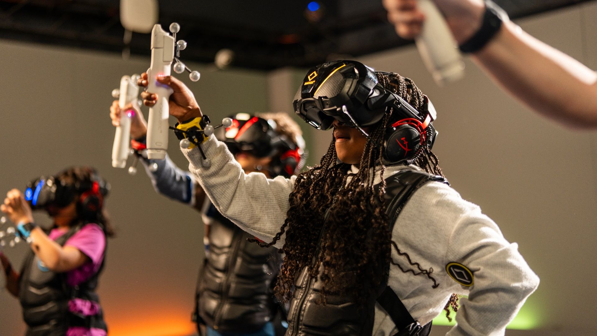 People in a VR arcade: foreground player with braided hair wearing a black headset and red headphones, raising white motion controllers; others in the background also wearing headsets.