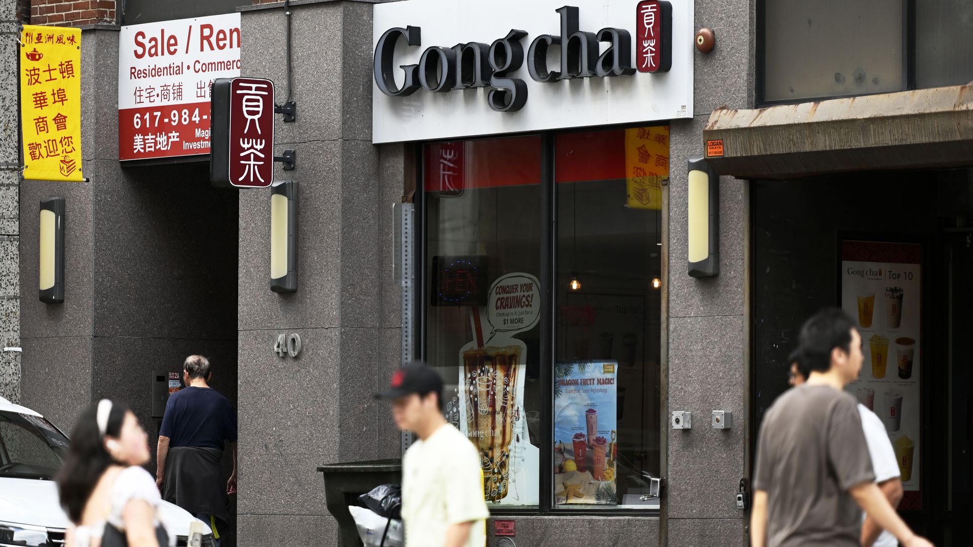 Exterior of Gong cha bubble tea shop with signs in English and Chinese, a red and white sale or rent sign, and several people walking on the sidewalk in front.