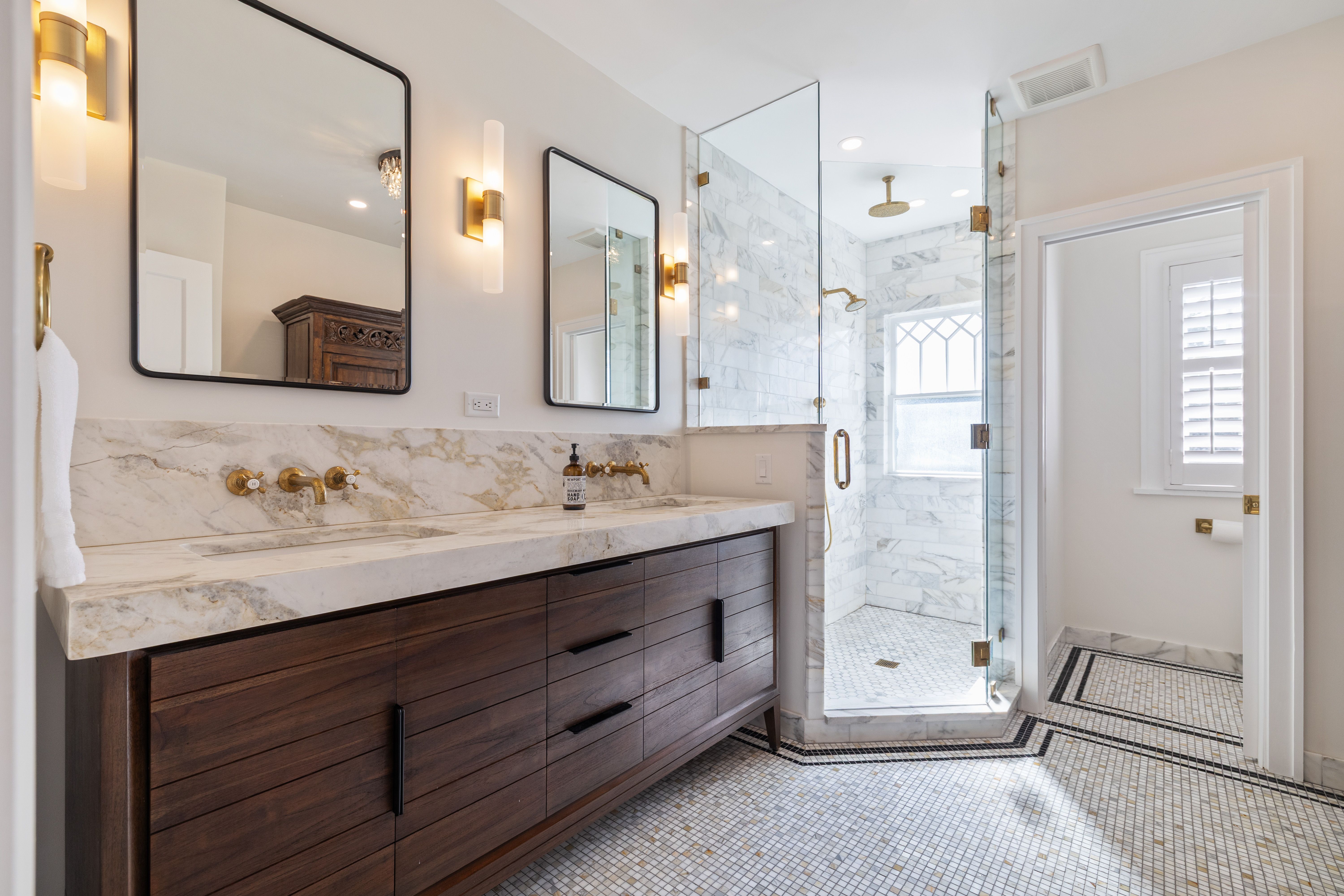 Modern bathroom with dark wood vanity, white marble countertop and backsplash, two rectangular mirrors, gold fixtures, glass shower with marble tiles, and mosaic tile floor.