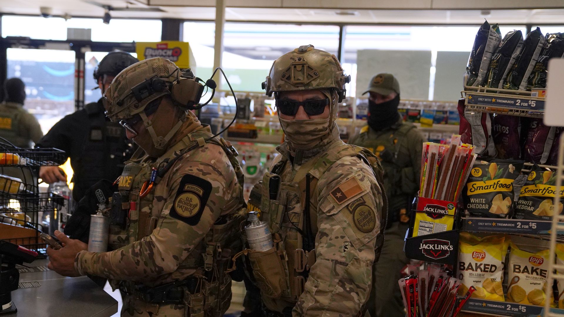 Armed law enforcement officers in tactical gear stand inside a convenience store, with shelves of snacks and drinks visible beside them.